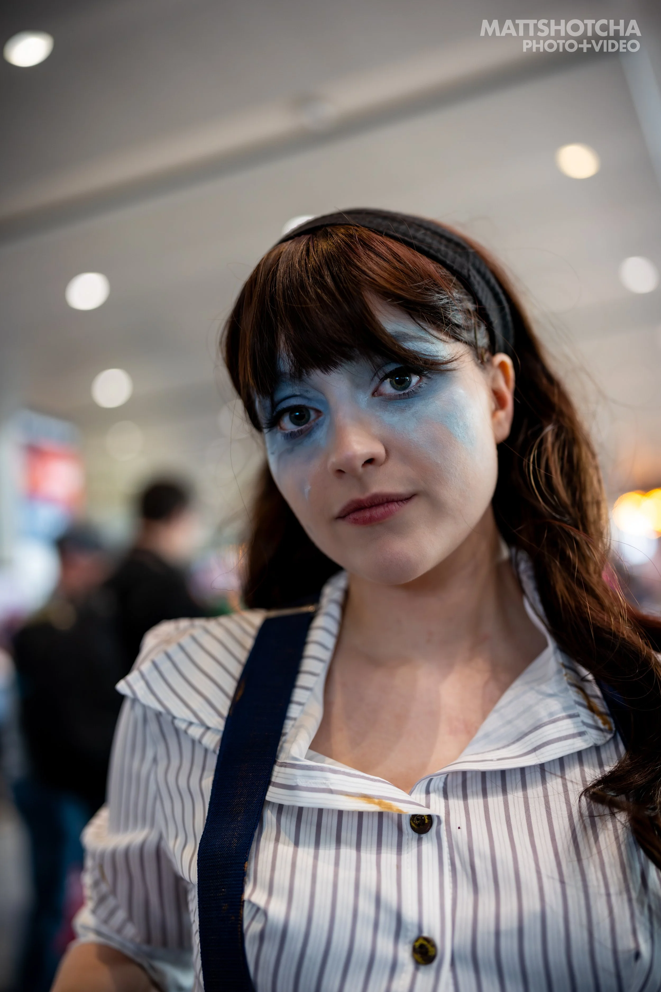 A young woman with brown hair and blue face paint around her eyes, wearing a striped shirt and a headband, standing in a busy indoor setting at PAX East 2026.