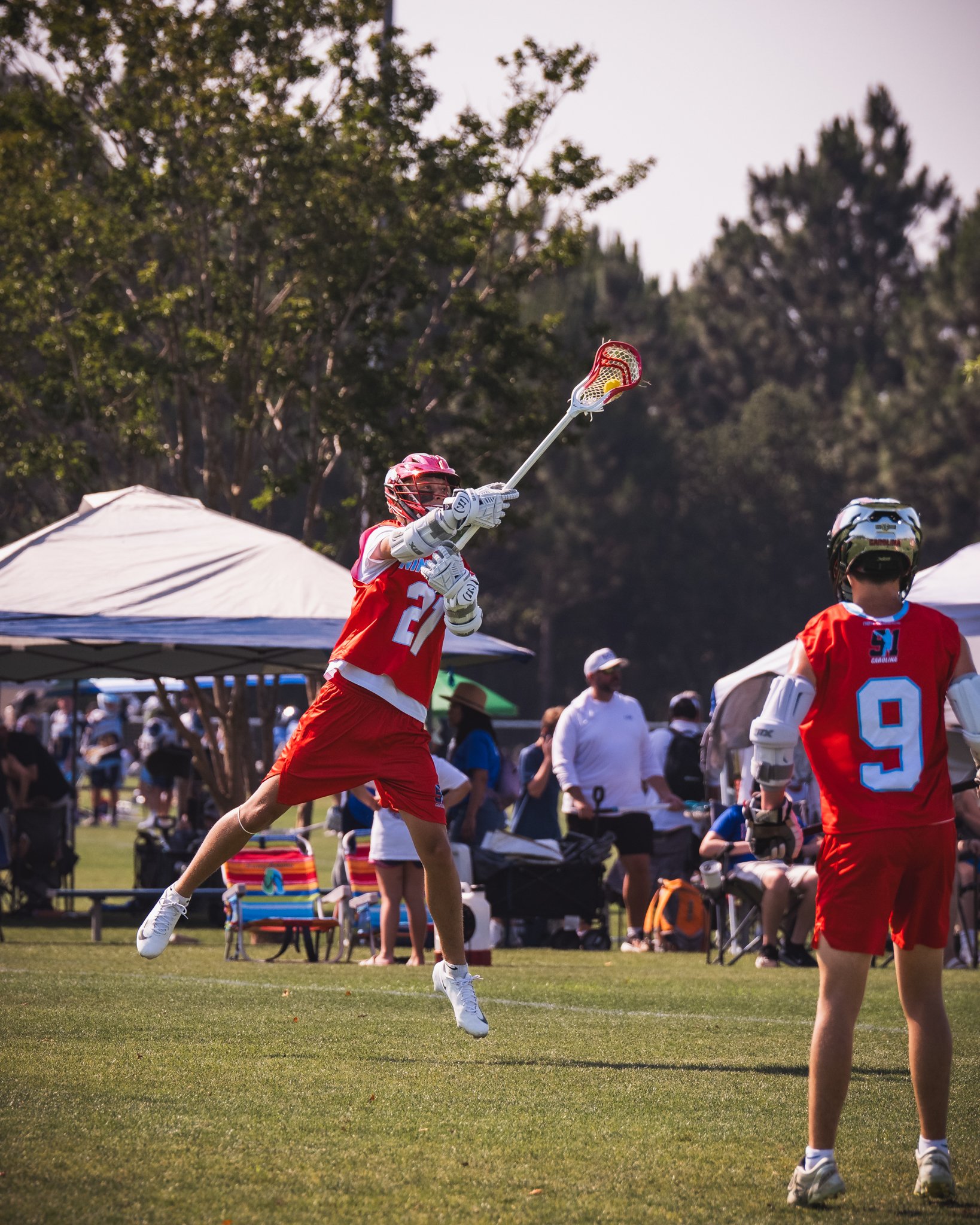 A Team 91 Carolina Lacrosse player, James Szep, wearing a red uniform and a helmet is jumping in the air, reaching with their lacrosse stick to catch or intercept a ball at an outdoor sports event.