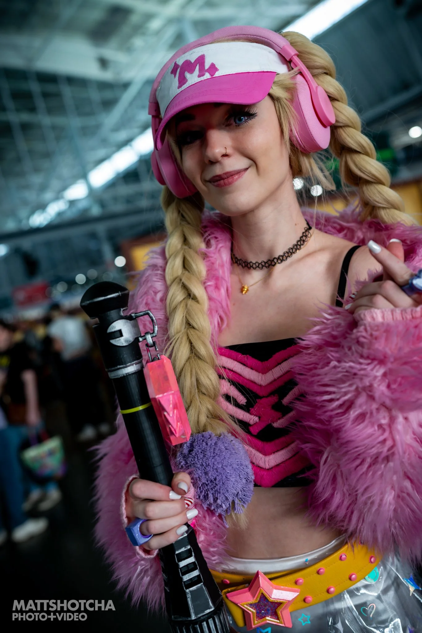 A woman, SuchKatie, dressed as New Millenia Might Magik from Marvel Rivals with pink fur coat, pink visor, and large pink headphones, holding a prop sword in a busy, brightly lit indoor space at PAX East 2026.
