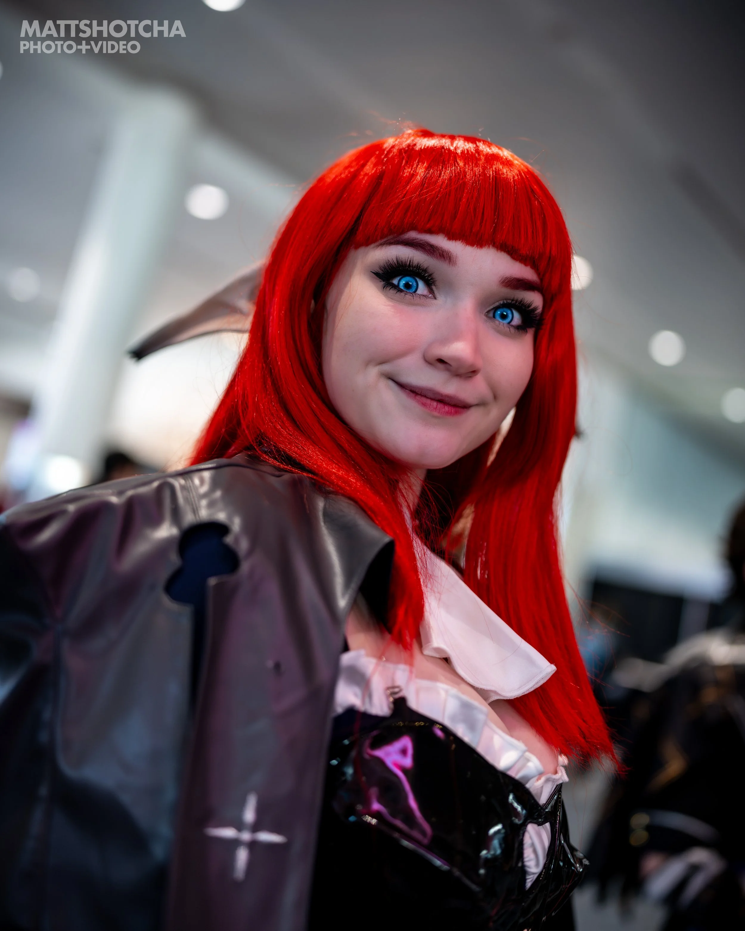 Close-up of a woman with bright red hair, blue eyes, and makeup, smiling at the camera, at PAX East 2026.