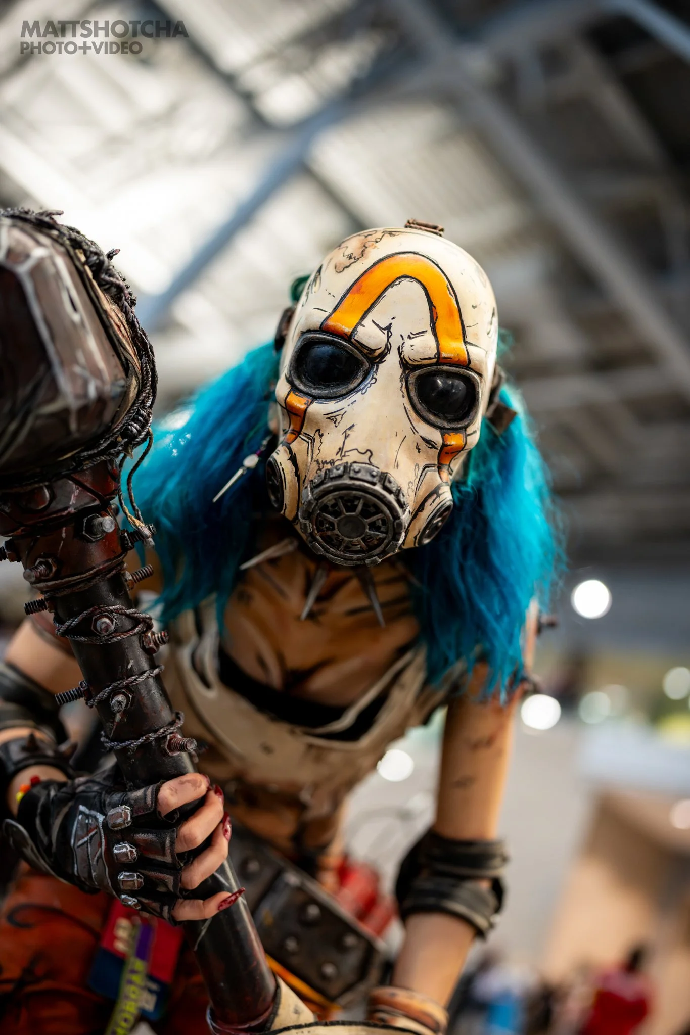 Close-up of a person wearing a Psycho mask from Borderlands with orange and black designs, blue hair, and cell shaded clothing, holding a weapon at PAX East 2026.