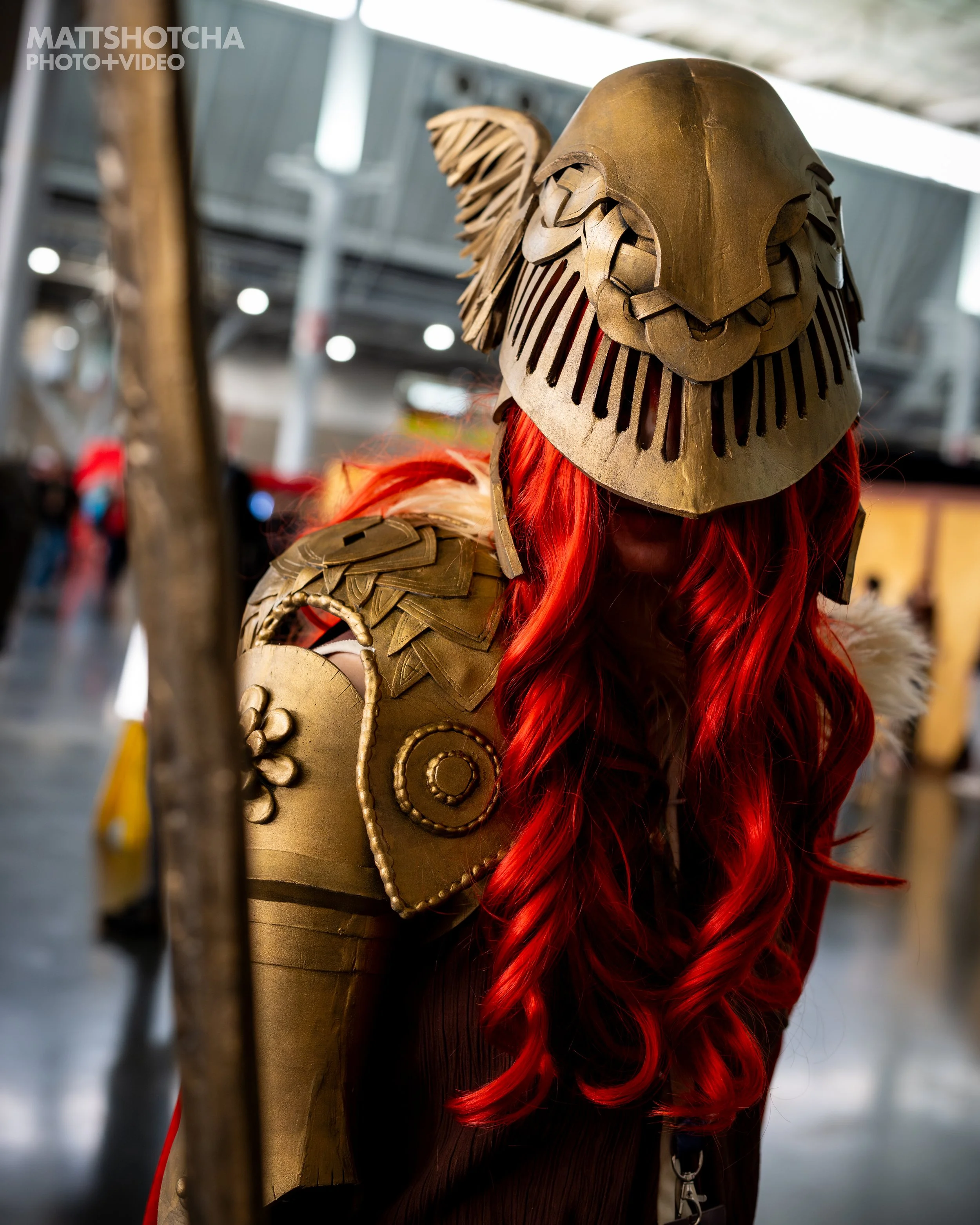 A person dressed in medieval knight armor with a helm and shoulder plates, with long red hair flowing out from beneath the helm, standing indoors with blurred background at PAX East 2026.