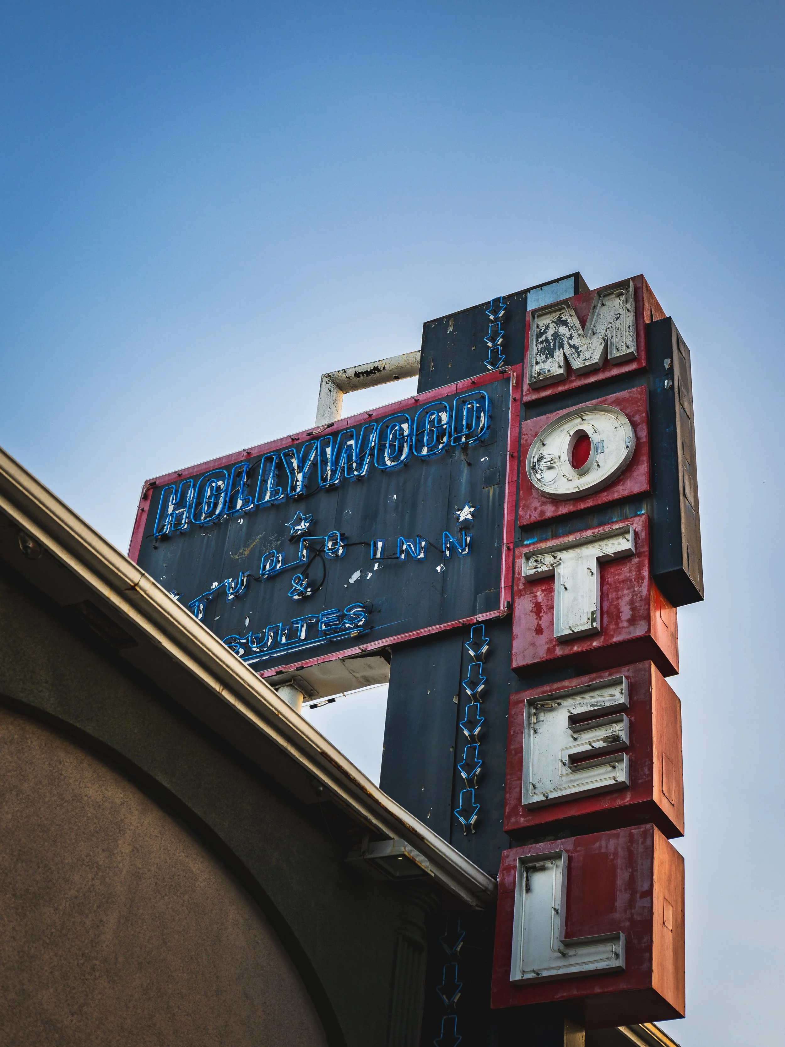 Close-up view of a retro neon sign with the word 'HOLEYWOOD' on top and various other words and symbols below, featuring broken and weathered signage on a building against a bright blue sky.