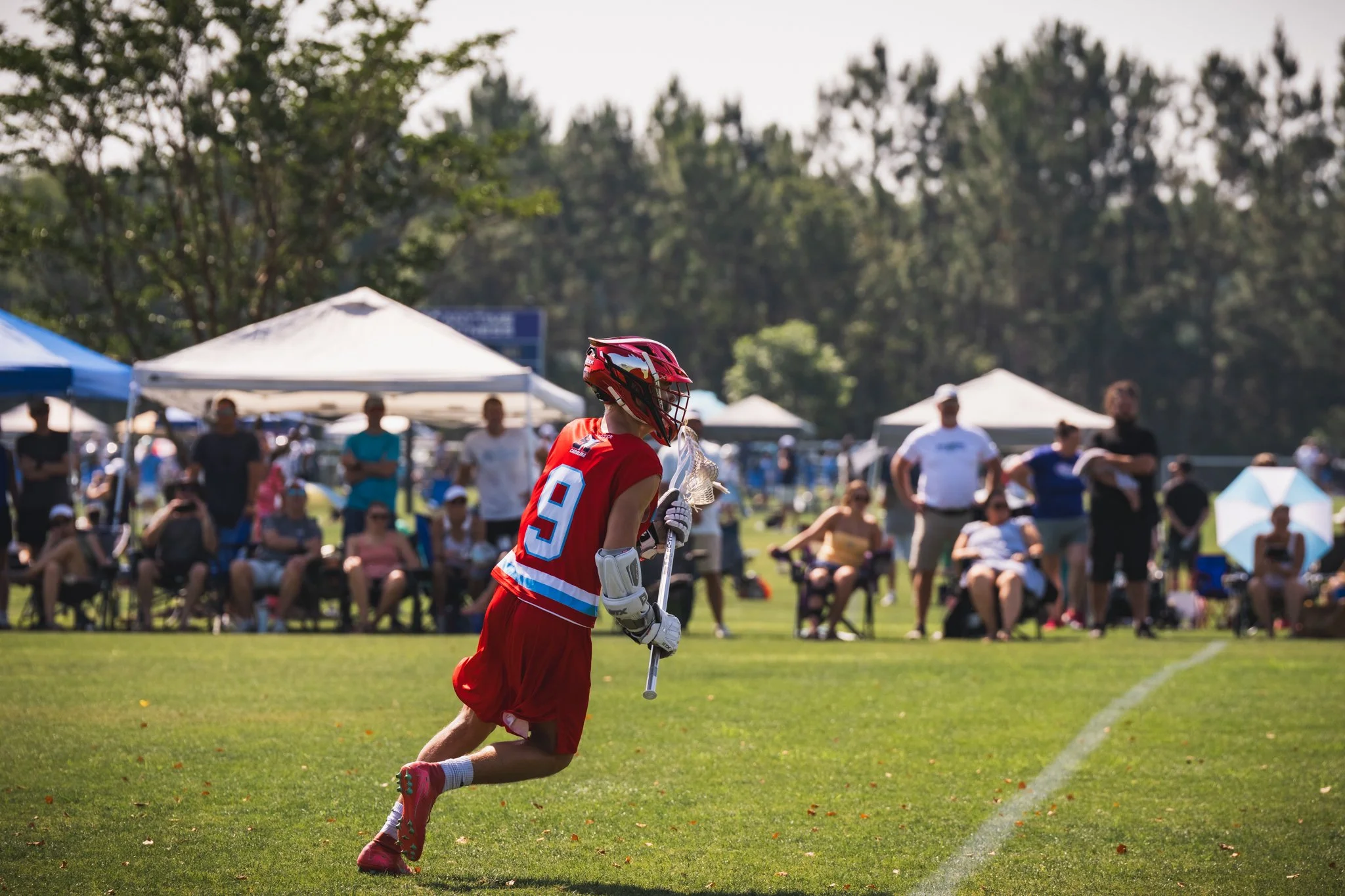 A Team 91 Carolina Lacrosse player in red uniform running on a grassy field surrounded by spectators and tents, with trees in the background.