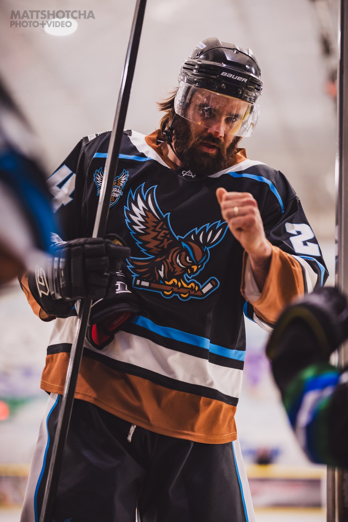 A Lexington Nightfall hockey player in a black jersey with a blue and brown owl logo, wearing a clear visor helmet, holding a hockey stick, and making a fist bump gesture.