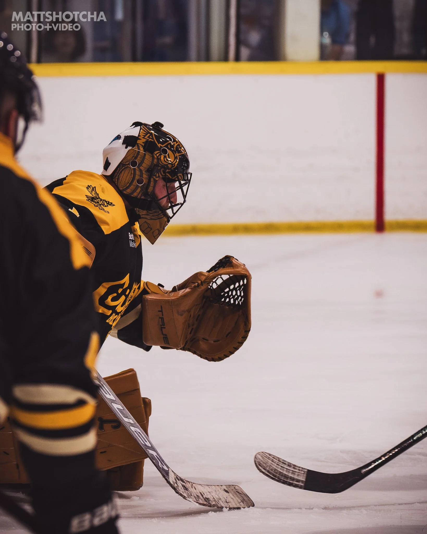 Lexington Nightfall goalie in yellow and black uniform and helmet, holding a brown goalie glove, on ice rink with hockey stick, with spectators in the background.