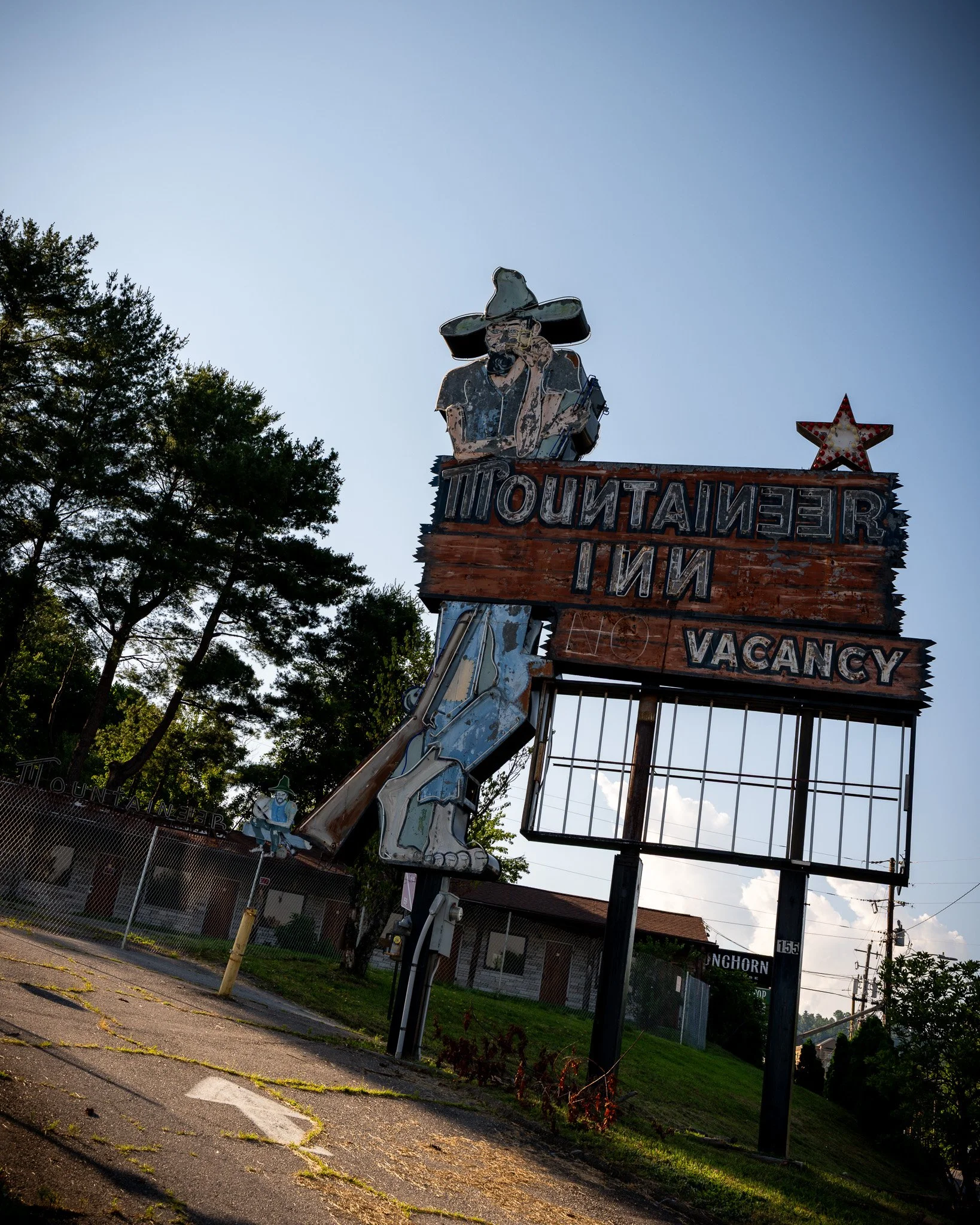 Old, weathered neon sign for a motel, featuring a figure of a man wearing a large hat and sunglasses, with the motel name The Mountaineer Inn visible, surrounded by trees and a chain-link fence.
