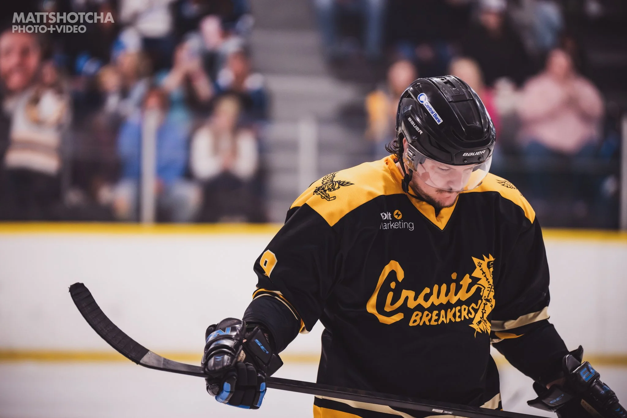 A Lexington Nightfall ice hockey player wearing a black and yellow jersey with the word "Circuit Breakers" and a logo, black helmet with a sticker for Central Bank, and holding a hockey stick during a game.