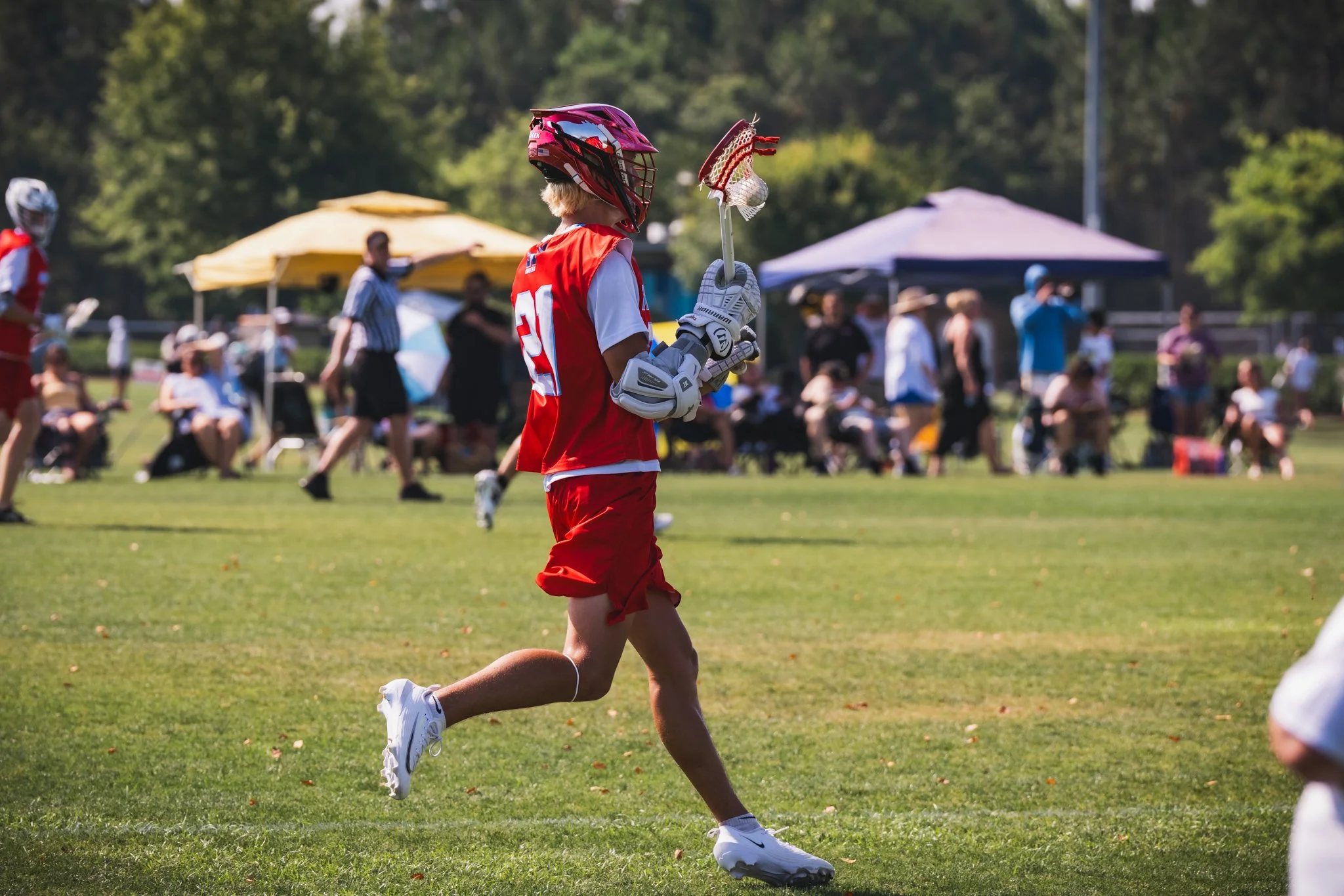 A Team 91 Carolina lacrosse player, James Szep, on a grassy field, wearing a red helmet, red uniform, and white shoes, holding a lacrosse stick with a ball in the net, with spectators and tents in the background.
