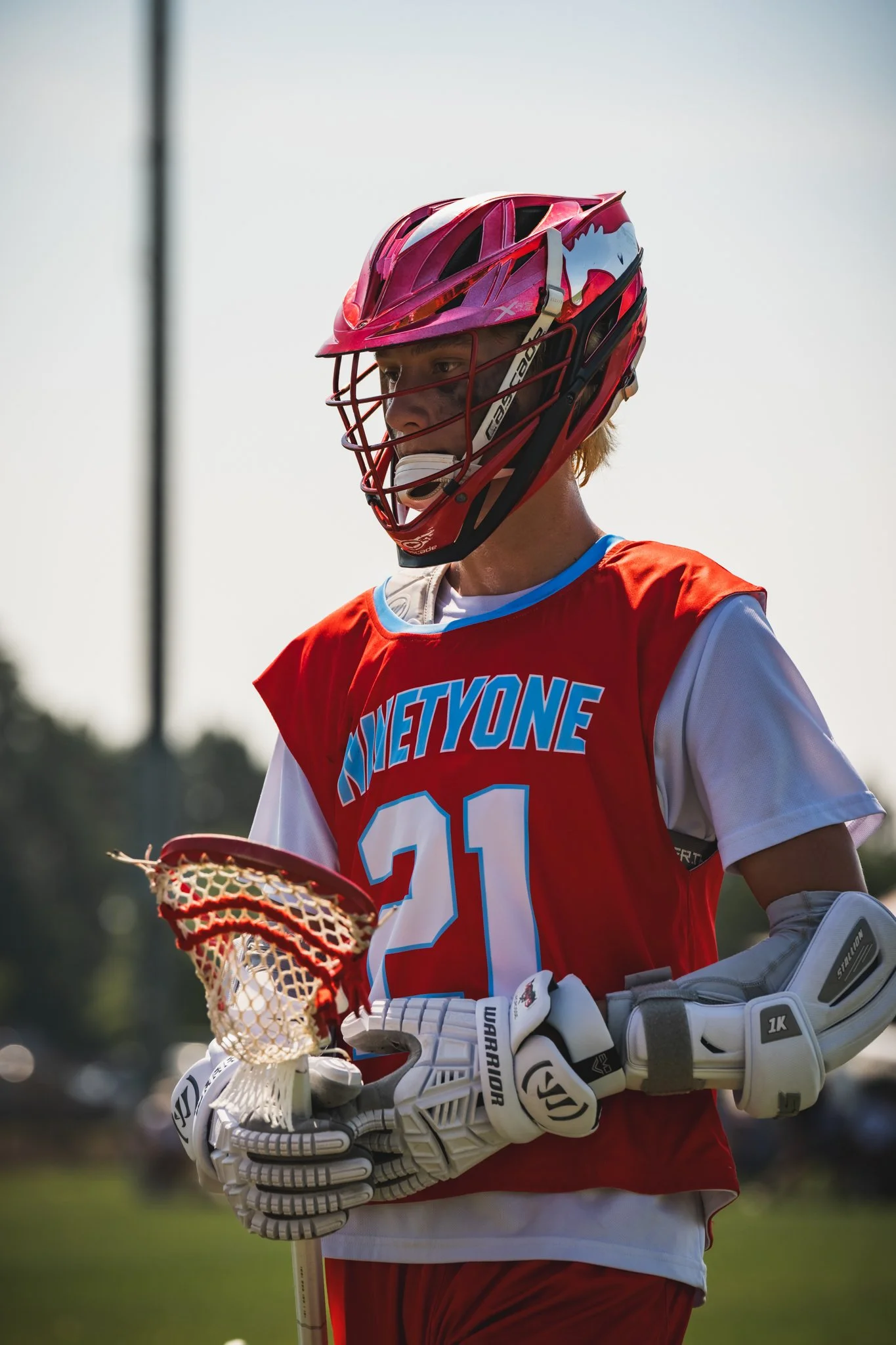 A Team 91 Carolina Lacrosse player, James Szep, wearing a red helmet and jersey with the number 21, holding a lacrosse stick, and standing outdoors on a sunny day.