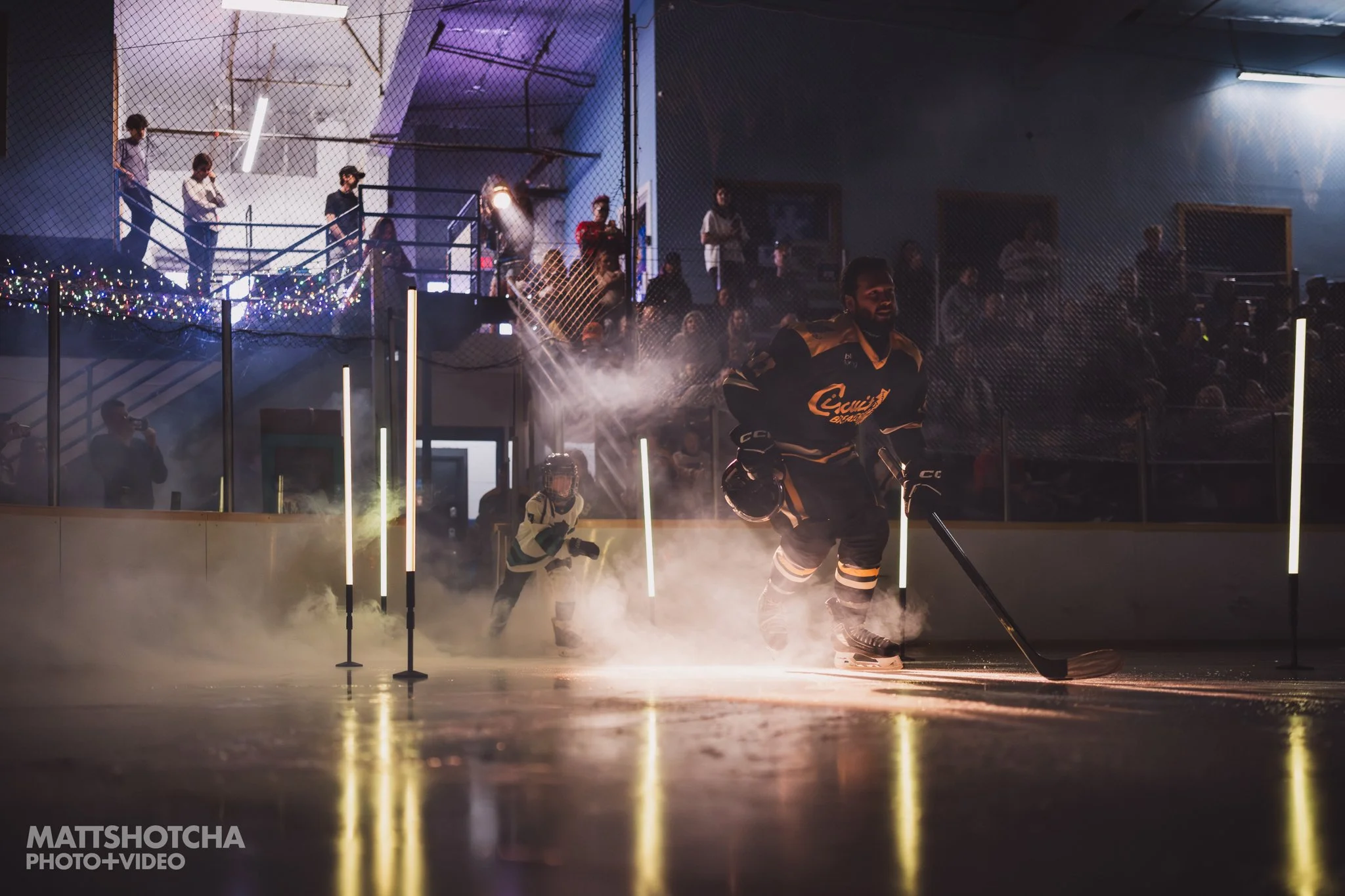Lexington Nightfall Hockey players on an ice rink with one player holding a hockey stick and younger players in the background, all surrounded by fog or vapor with bright lights and a crowd of spectators in the background.