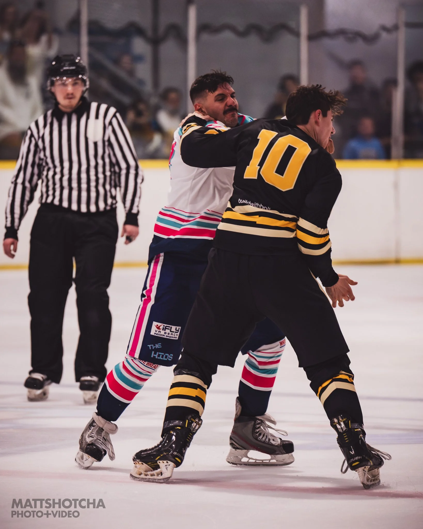 Two hockey players engaged in a fight on the ice rink during a Lexington Nightfall game, with a referee nearby. One player is wearing a black and yellow jersey, the other a colorful jersey with stripes. Spectators are blurred in the background.