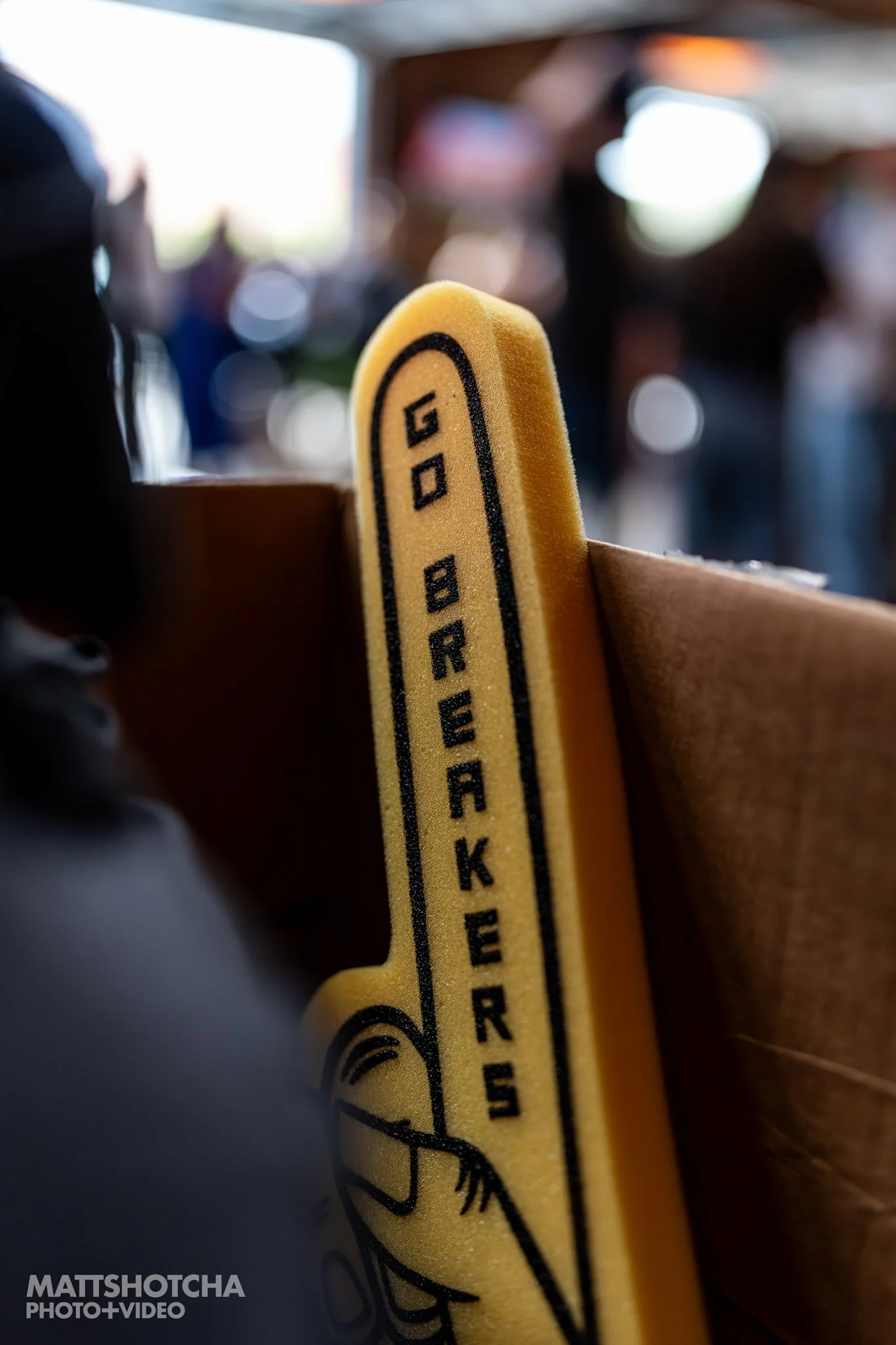 Close-up of a yellow foam finger with black text and illustration, placed in a wooden holder, with blurred background of people at a social event.
