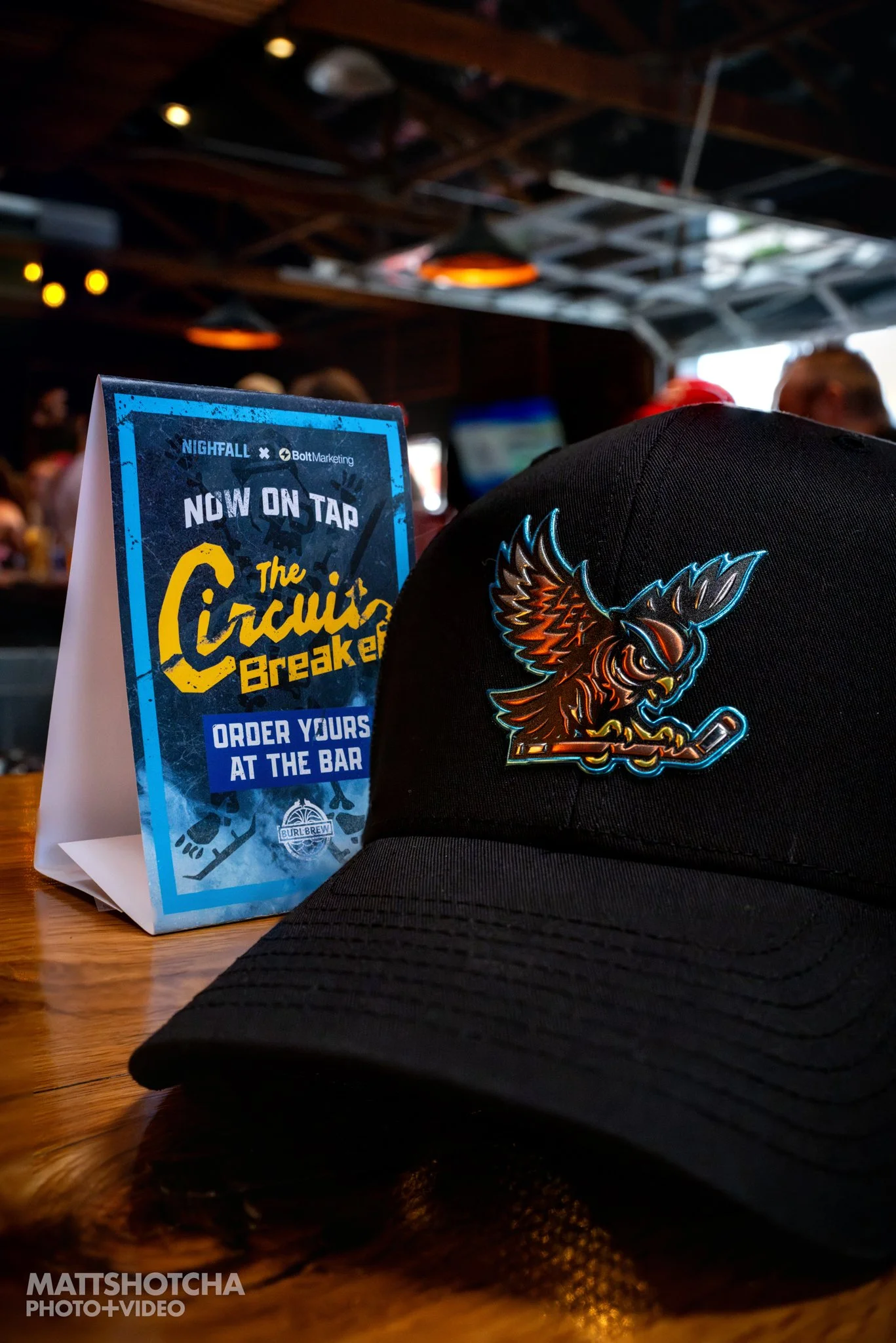 A black cap with a colorful embroidered Lexington Nightfall owl on the front, placed on a wooden bar. To the left, a blue promotional sign is visible. The setting is The Burl in Lexington Kentucky.