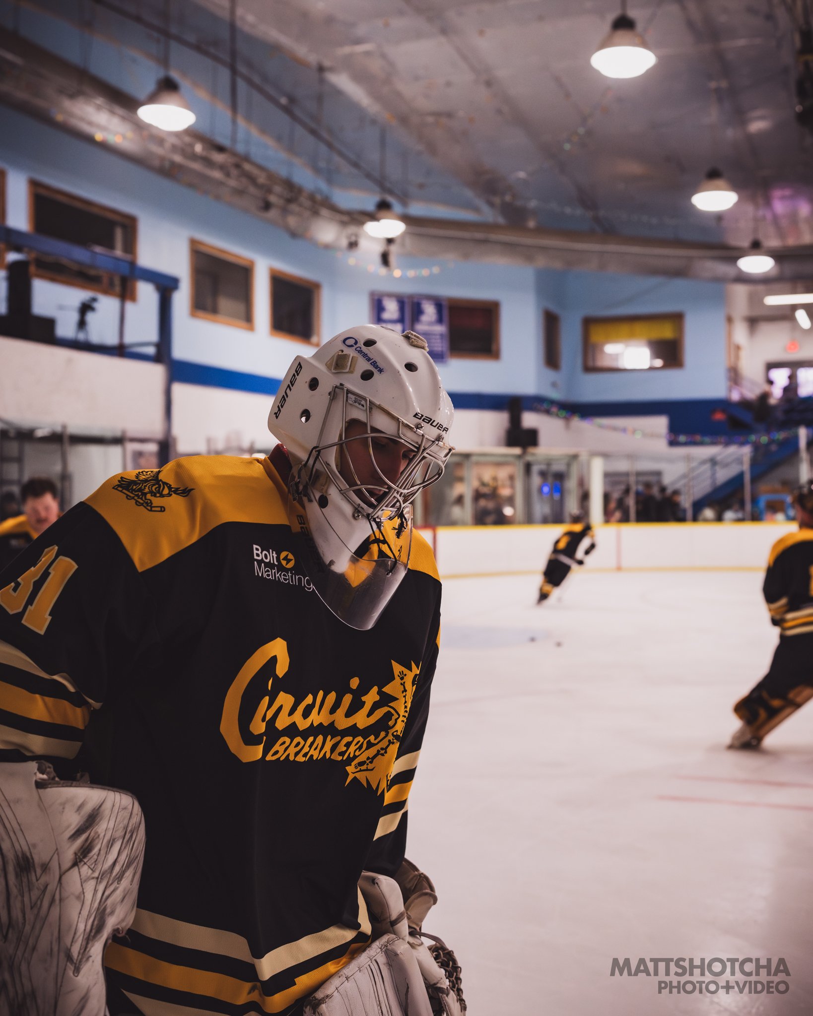 A Lexington Nightfall ice hockey goalie wearing a yellow and black jersey with the words "Circuit Breakers" on it, a white helmet with a face shield, in an indoor ice rink. Other players are in the background.