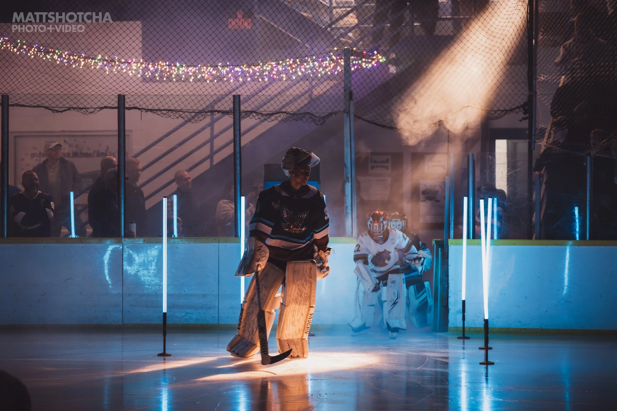 An indoor ice hockey rink with players and a goaltender for the Lexington Nightfall, illuminated by colorful string lights and a spotlight, with spectators watching behind the protective glass.