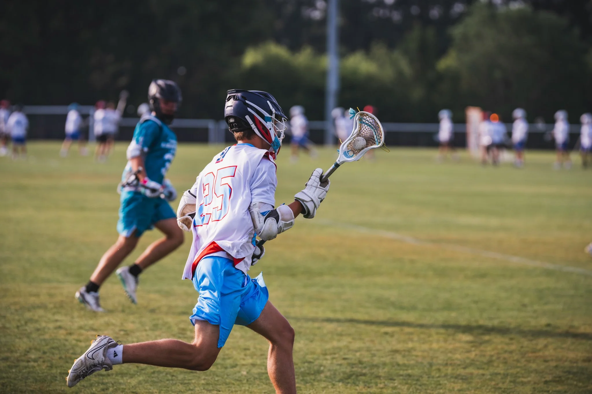 A Team 91 Carolina Lacrosse player in white and red uniform running with lacrosse stick on field, with other players in background.