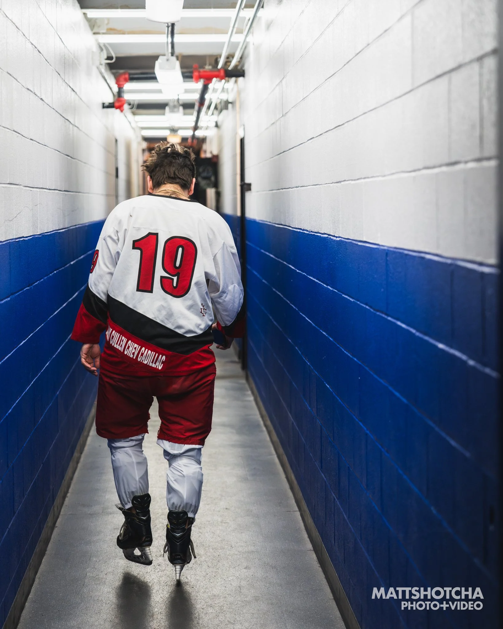 A person with short dark hair wearing a white hockey jersey with the number 19 on the back, black ice skates, and striped white hockey pants,  walking down a narrow corridor with blue walls and exposed ceiling pipes.