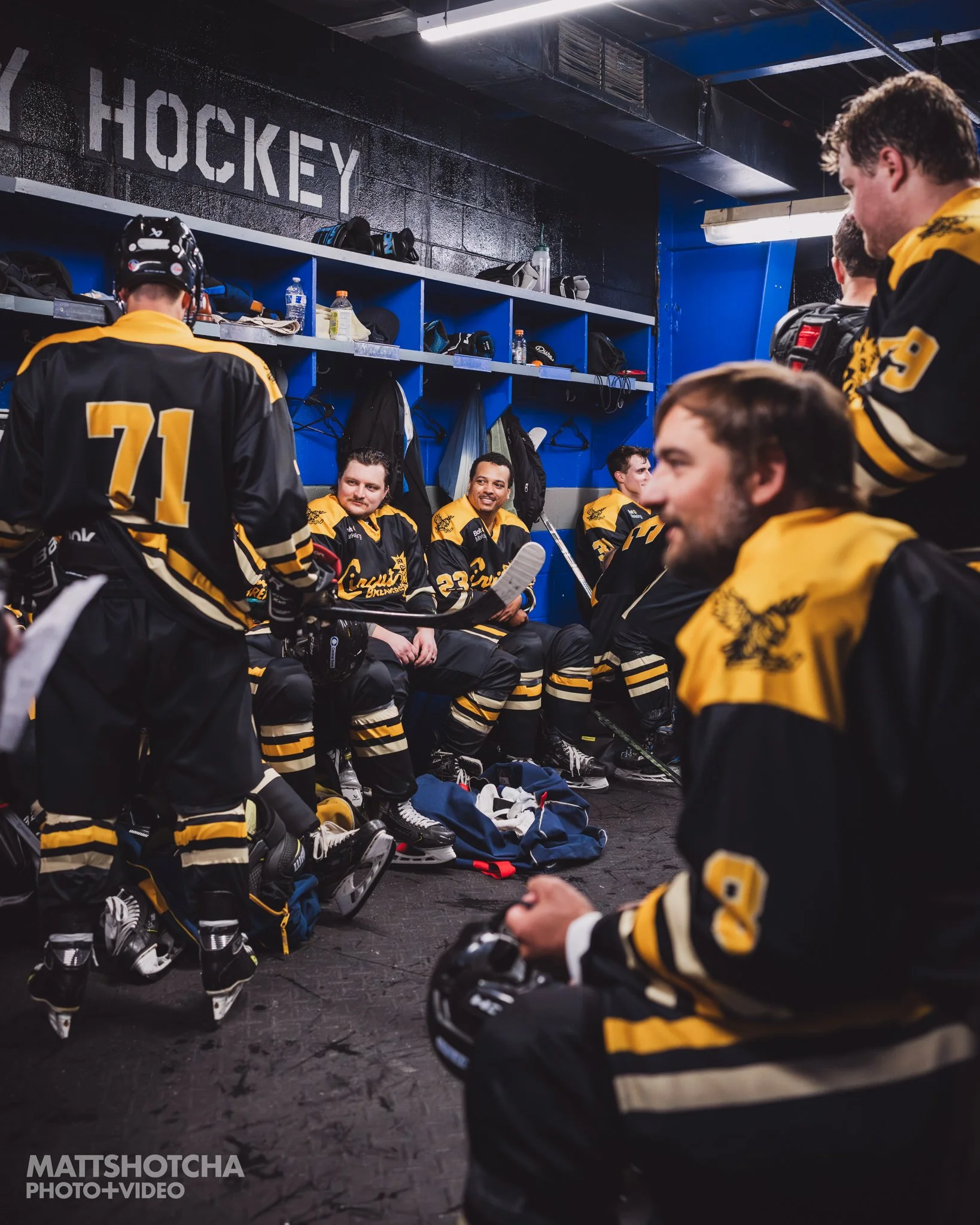 Lexington Nightfall hockey players sitting and standing in a locker room, some with helmets and skates, wearing black and yellow uniforms, near blue lockers with gear and water bottles.