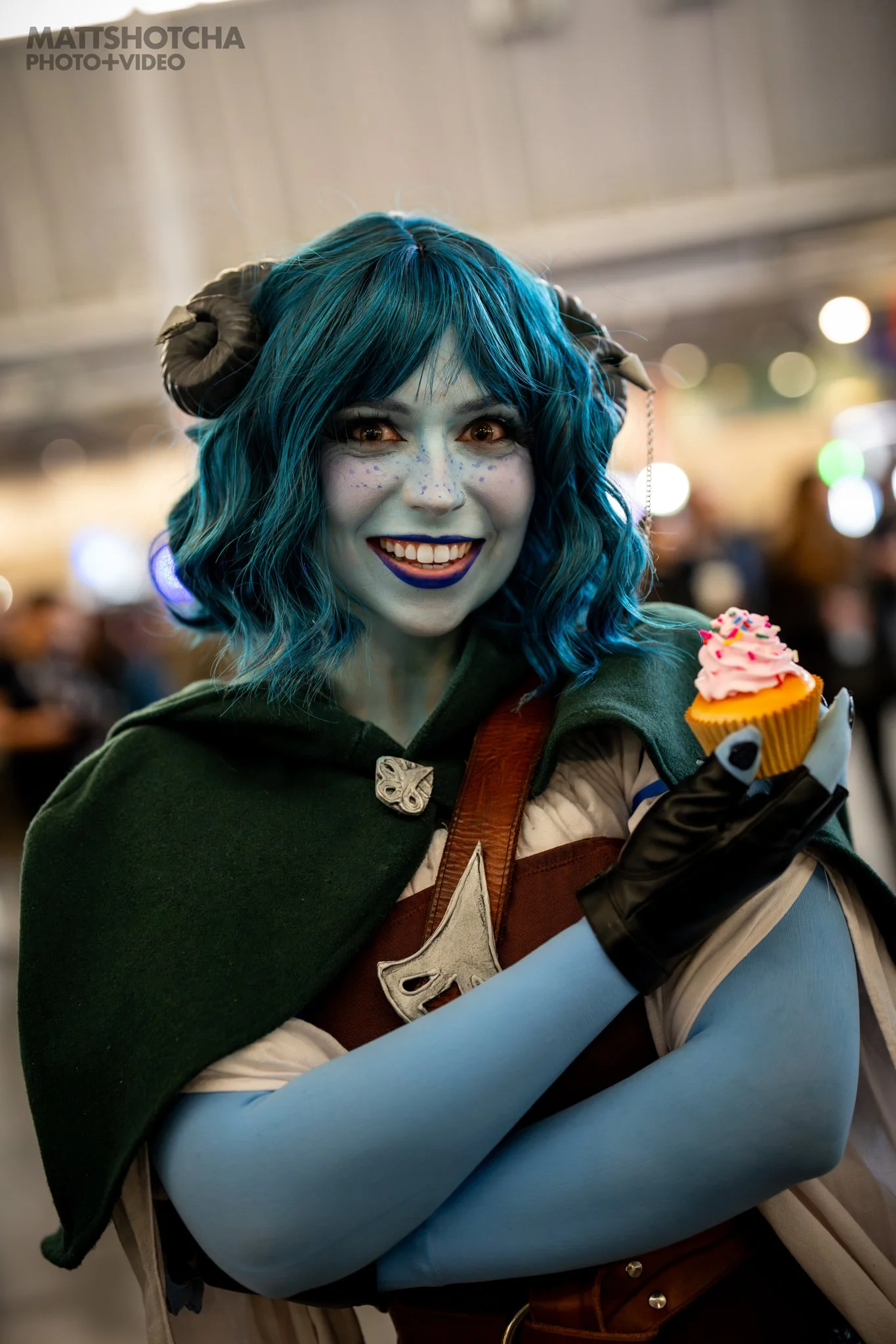 A woman, BostonBeeCosplay, dressed as a character from Critical Role with blue hair and makeup holding a cupcake with pink frosting and sprinkles, smiling, at PAX East 2026.