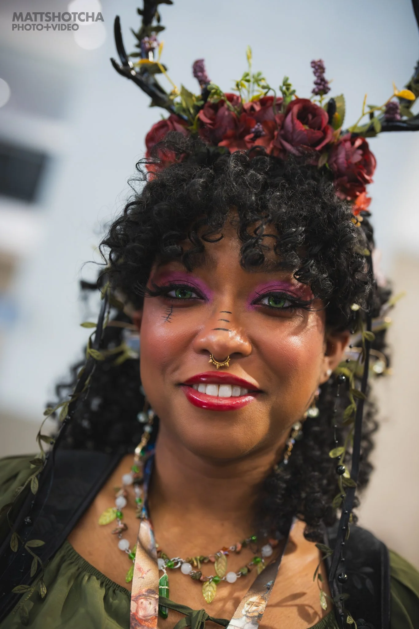 A woman with curly black hair and bright green eyes smiling, wearing colorful makeup with pink eyeshadow and red lipstick, and adorned with a floral headpiece and jewelry.