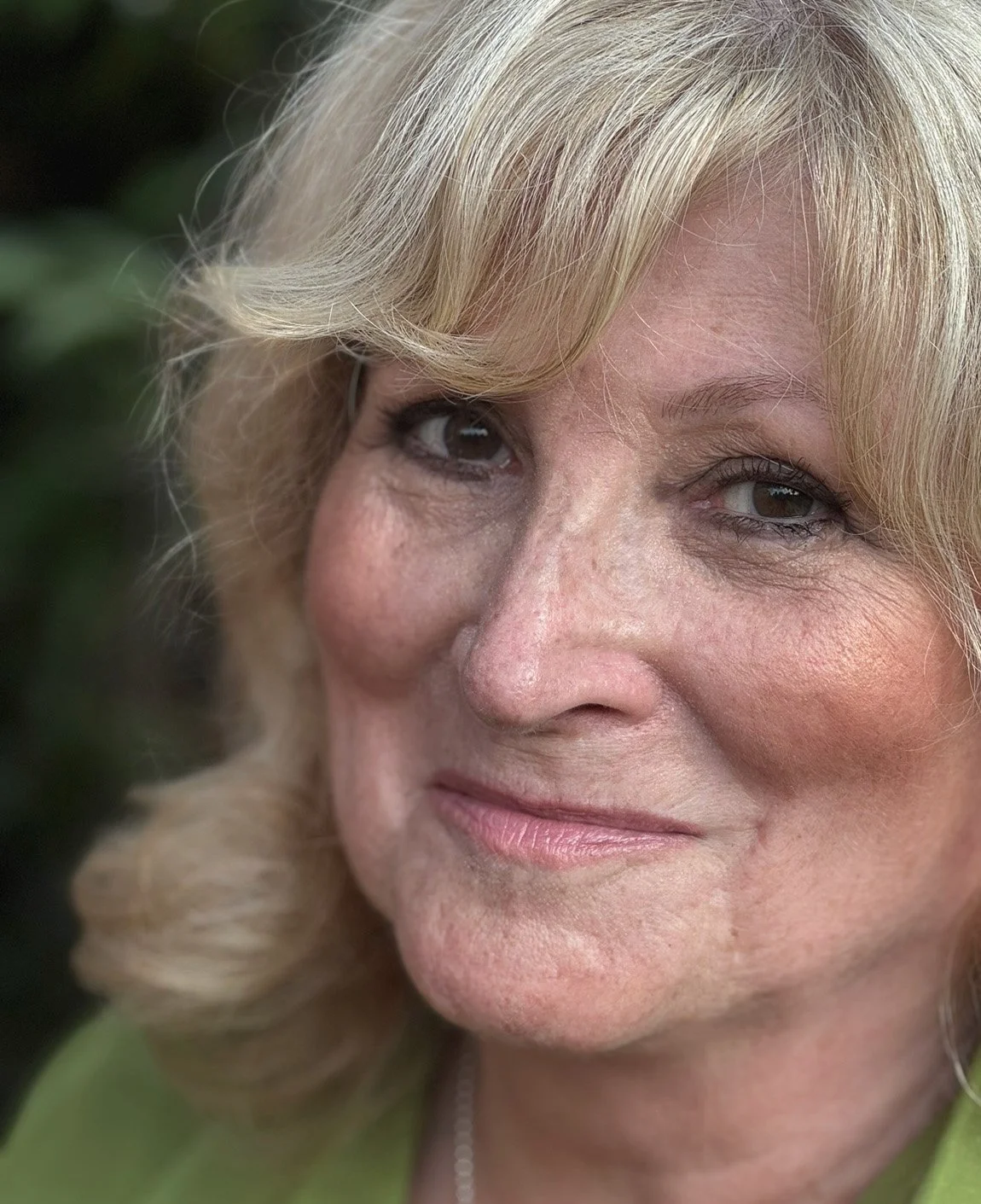 Close-up of a middle-aged woman with blonde hair, smiling, in front of a blurred background of green leaves