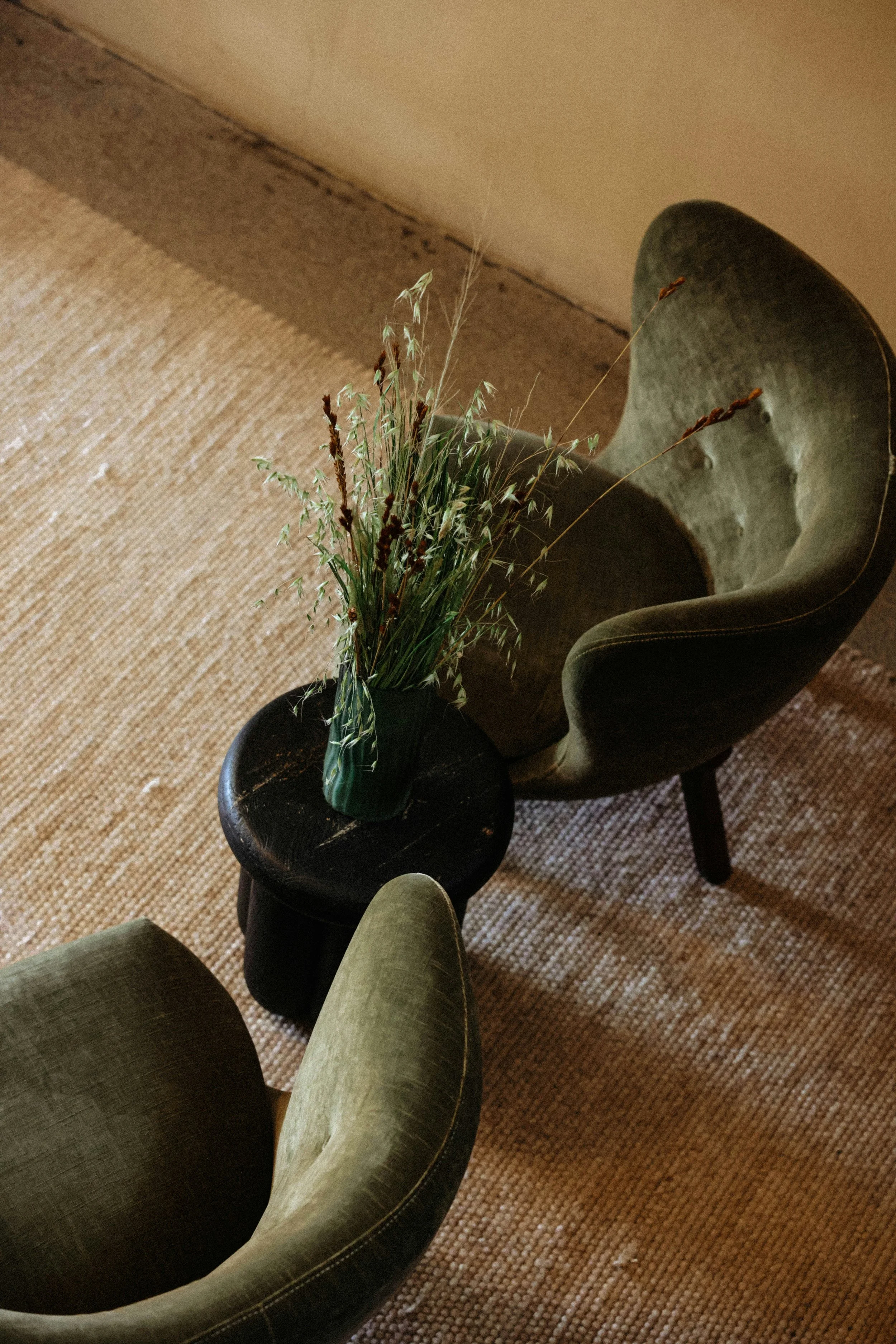 Vista de uma sala com uma cadeira de veludo verde, uma mesa menor de madeira escura com vaso de flores secas e um tapete de sisal.
