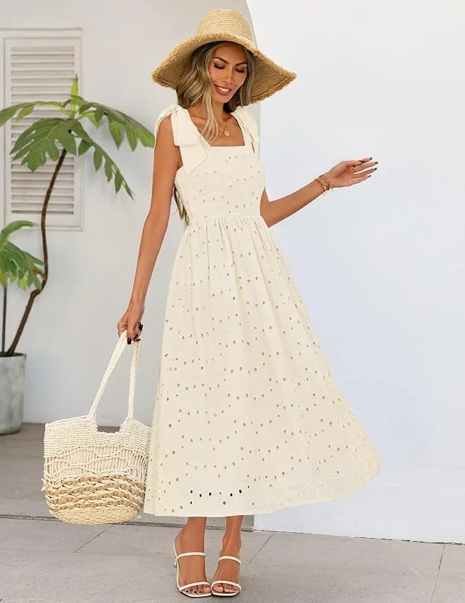 A woman wearing a white, eyelet maxi dress with a square neckline, wide shoulder straps, and a gathered waist, accessorized with a large straw hat, white high-heeled sandals, and holding a woven straw tote bag. She is standing against a white wall with a potted plant nearby.