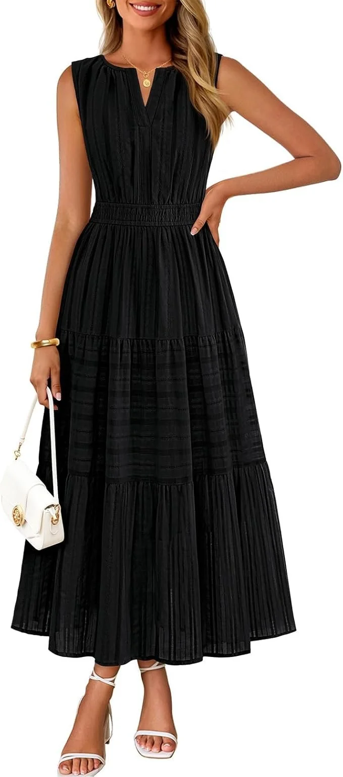 A woman wearing a sleeveless black pleated maxi dress, white high-heeled sandals, a gold necklace, and a white handbag, posing with one hand on her hip.