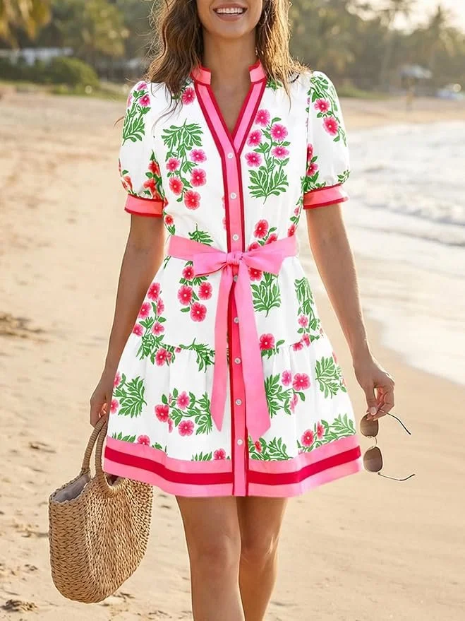 Woman in a floral dress walking on the beach, holding a straw bag and sunglasses.