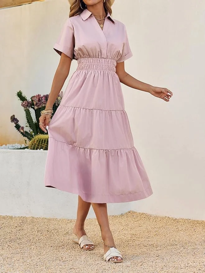 Woman wearing a pink, tiered, midi-length dress with short sleeves, standing on a gravel surface near a white wall with potted cacti and flowers in the background.