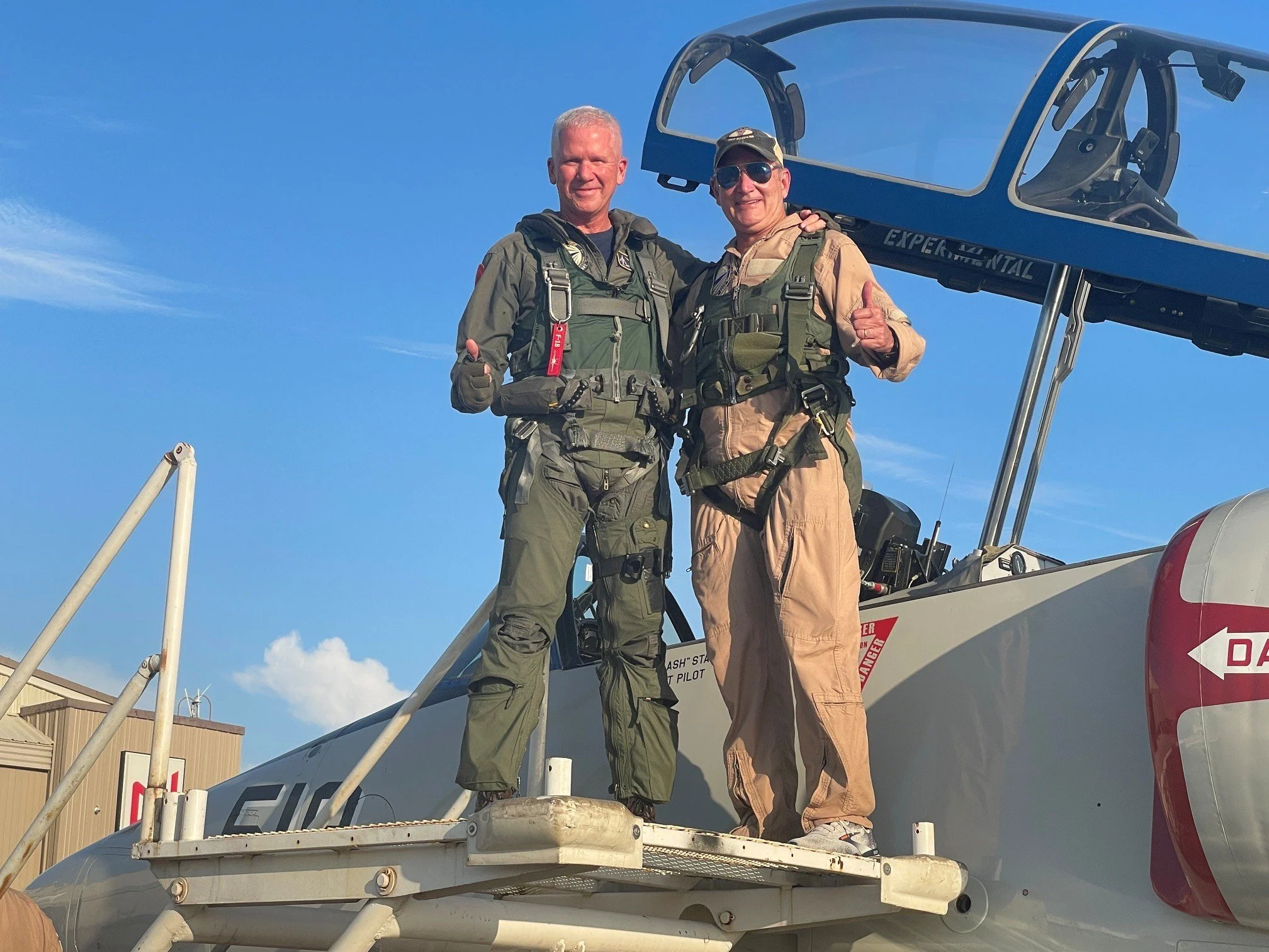 Two men in pilot uniforms standing on the step of a small aircraft, smiling and giving thumbs up, with a clear blue sky and airport buildings in the background.