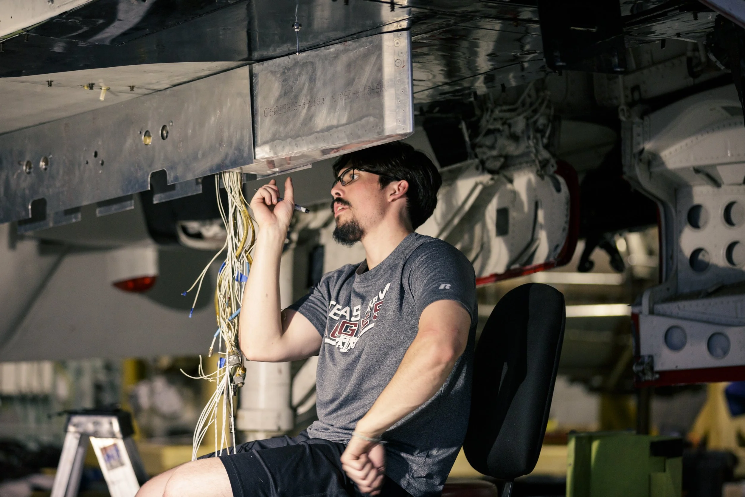 A man with glasses and a beard sitting under a spacecraft, working with wires and electronic components.