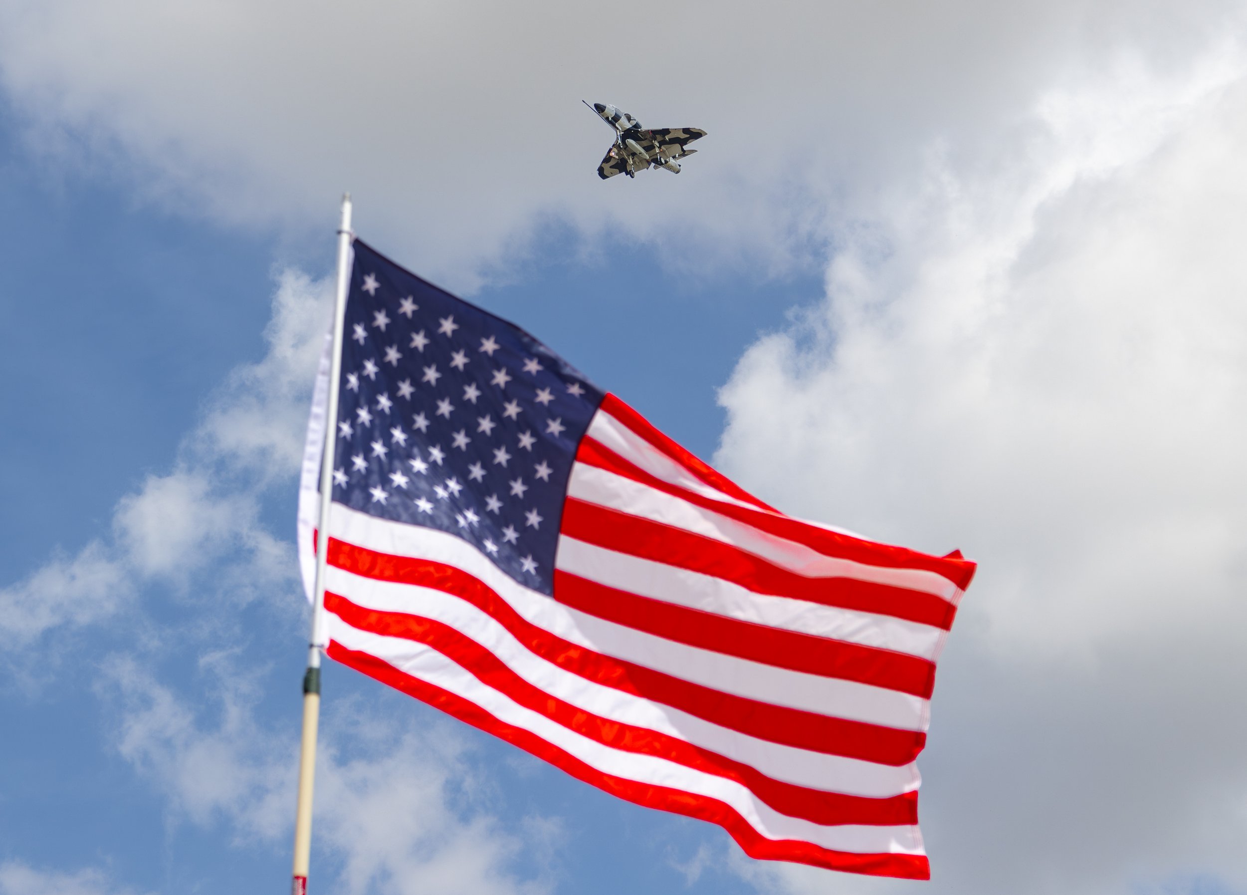 American flag waving in the wind with a military jet flying overhead through a partly cloudy sky.