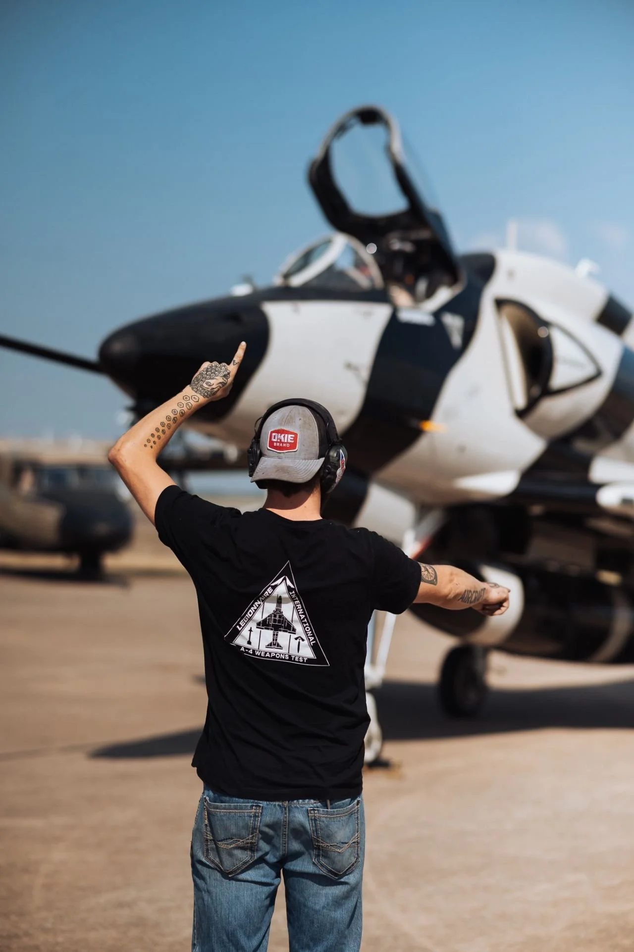 A person with tattoos on their arm, wearing a black t-shirt and a cap, signals with one hand in front of a fighter jet on an airstrip.