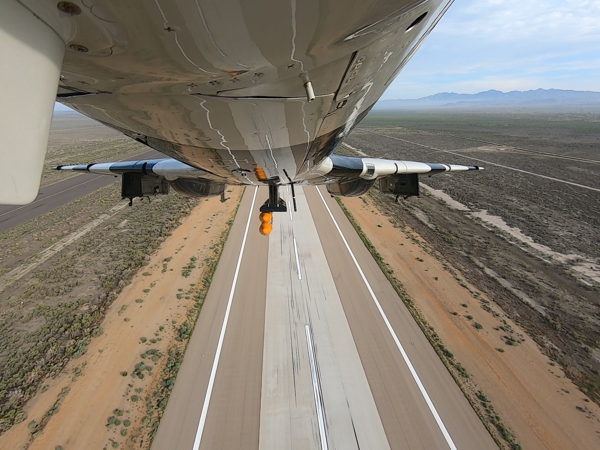 Underneath view of a jet flying over a desert with a clear sky and mountains in the background.