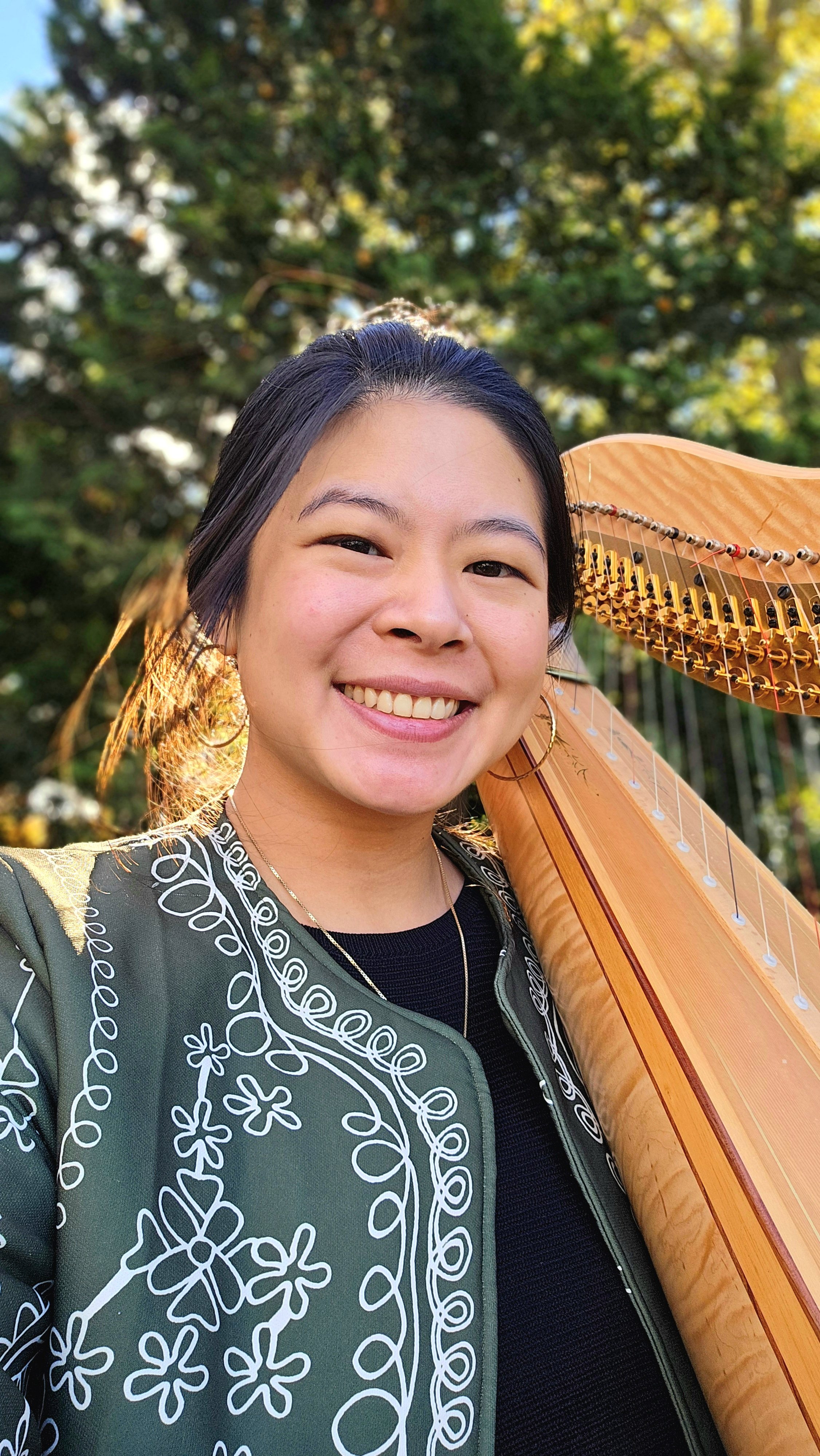 A smiling young woman with black hair and large hoop earrings, holding a wooden harp, standing outdoors with green trees and sunlight in the background.