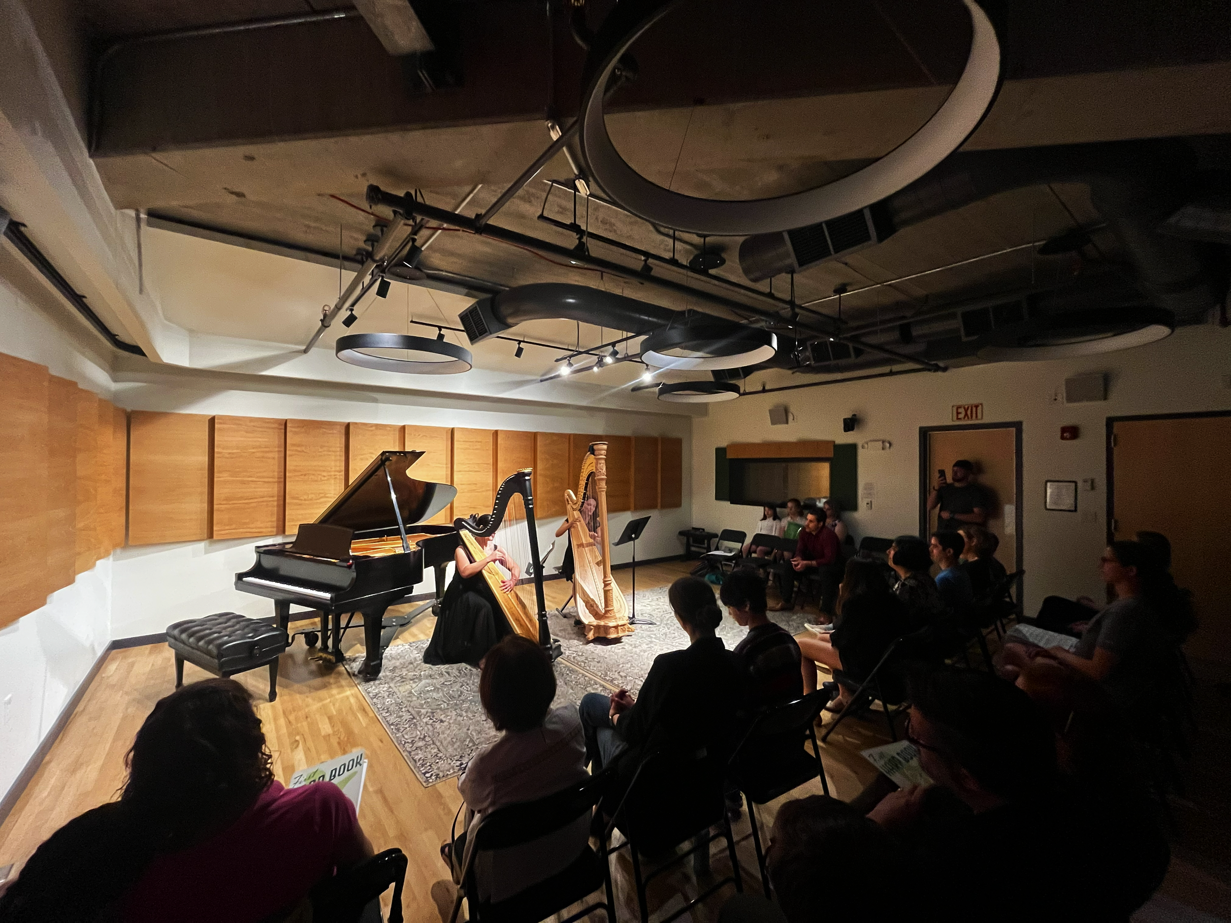 A musical performance featuring a person playing a harp and another playing a grand piano in a room filled with seated audience members, with microphones and stage lighting overhead.