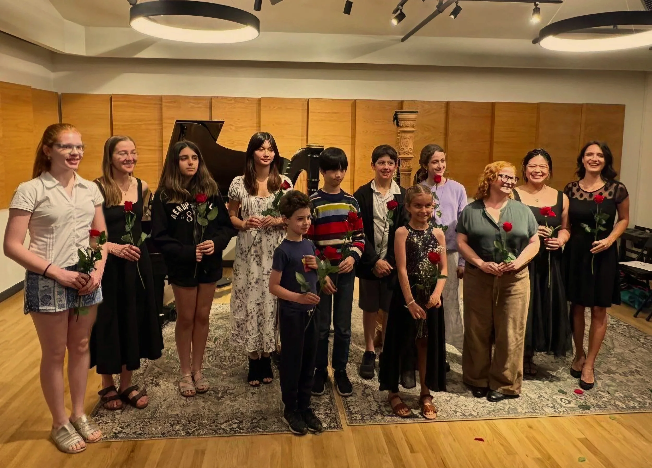 A group of people, including children and adults, are standing together holding red roses in an indoor setting with wooden walls and a grand piano in the background. The group appears to be participating in a celebration or concert.
