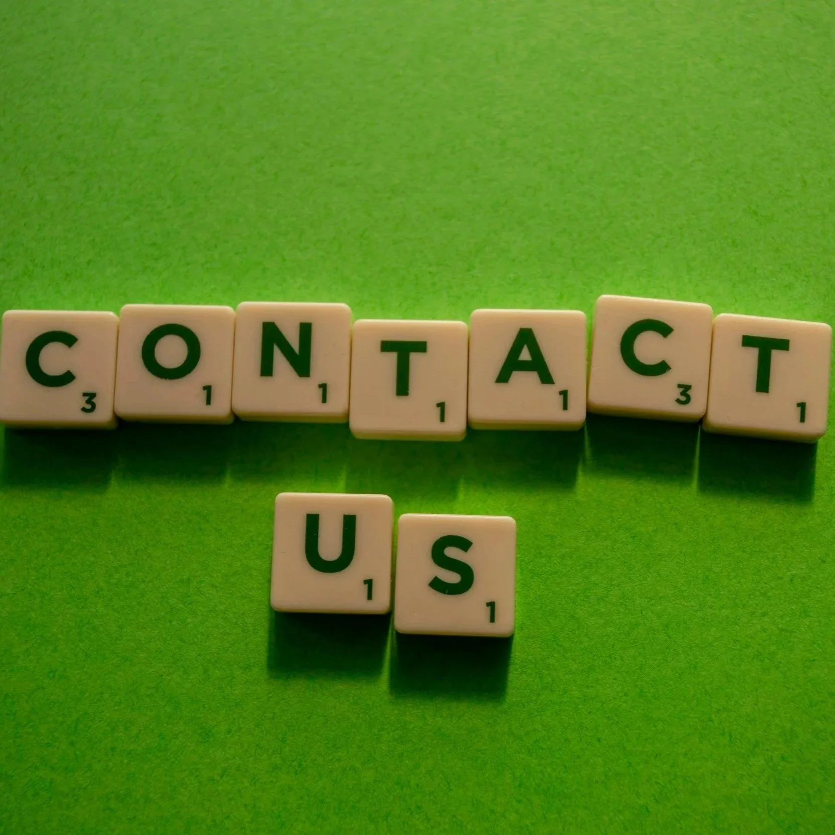 Scrabble tiles spelling the word 'CONTACT' on a green surface with additional tiles showing 'US' and 'I' below.