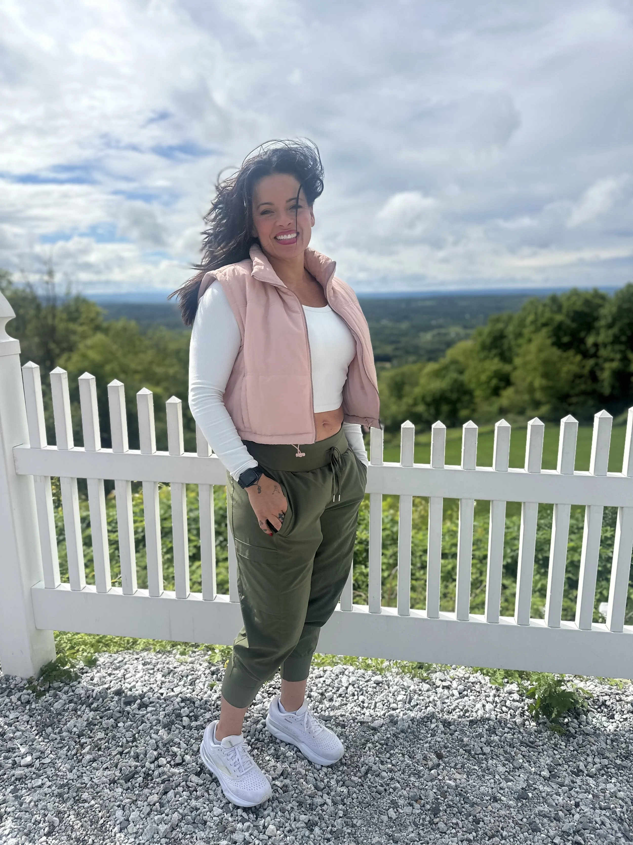 A woman with long black hair smiling, wearing a pink vest, white long sleeve shirt, olive green pants, and white sneakers, standing outside near a white fence with natural landscape and cloudy sky in the background.