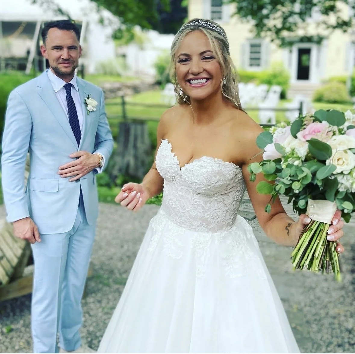 A bride holding a bouquet of flowers, smiling, with a man in a light blue suit standing outdoors in a garden area.