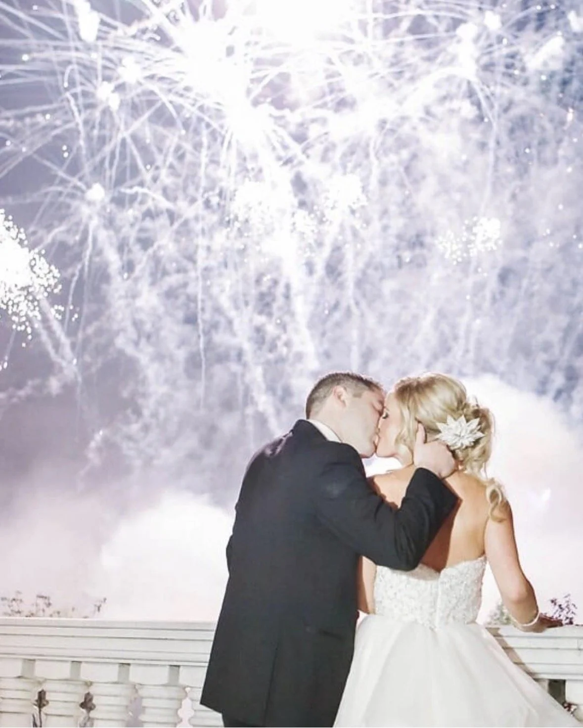 A bride and groom kissing during their wedding celebration with fireworks in the background.