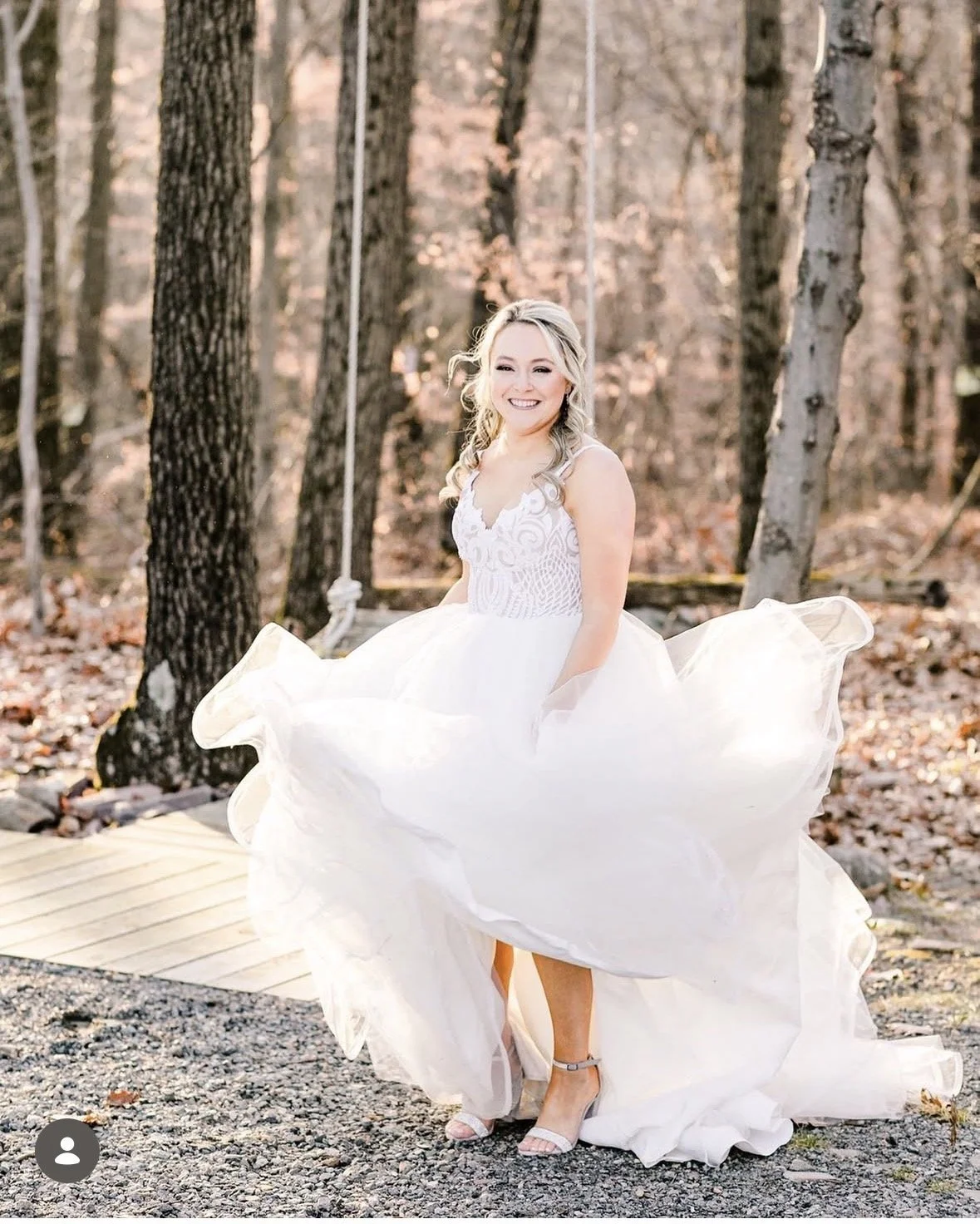 A young woman in a white wedding dress smiling outdoors in a wooded area, standing on a wooden boardwalk.
