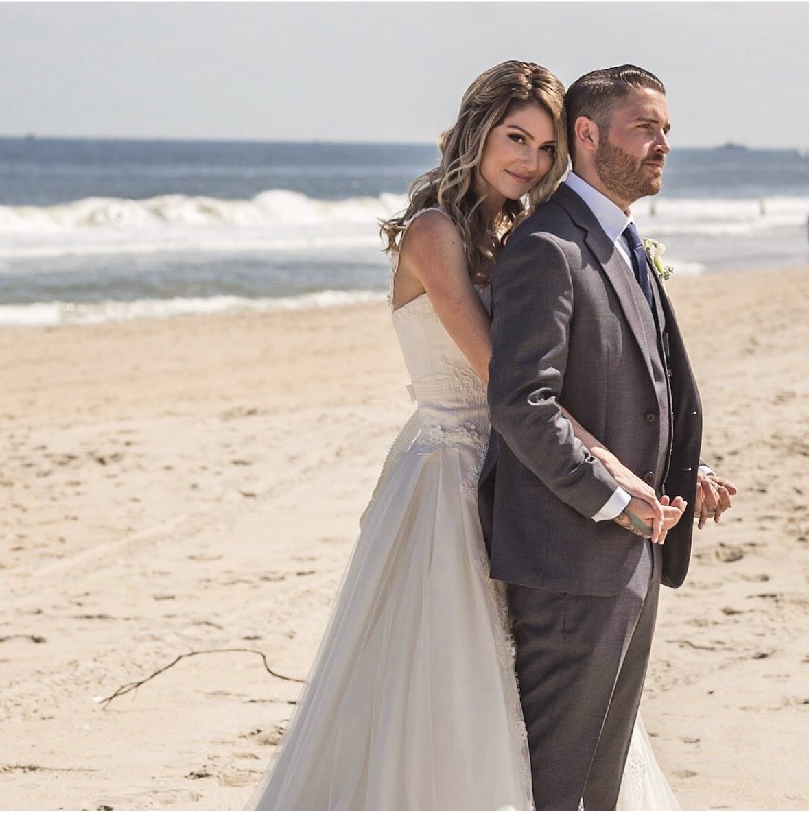 A bride and groom stand on a sandy beach during their wedding, holding hands, with ocean waves in the background.