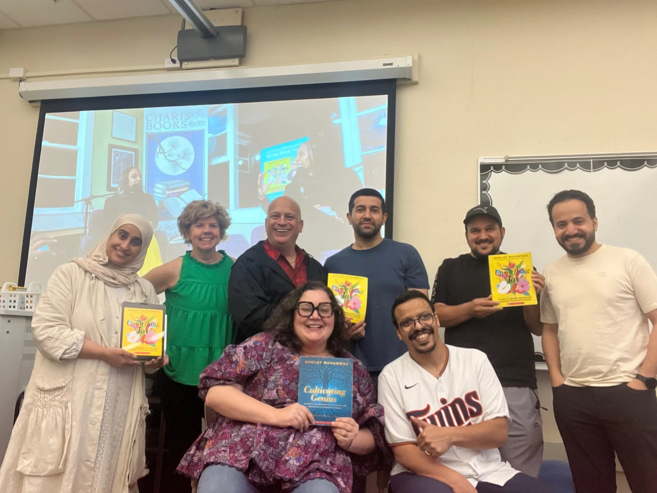 Group of diverse people, some holding colorful books, smiling and posing for a photo in a classroom, with a projection screen showing a bookshelf and books behind them.