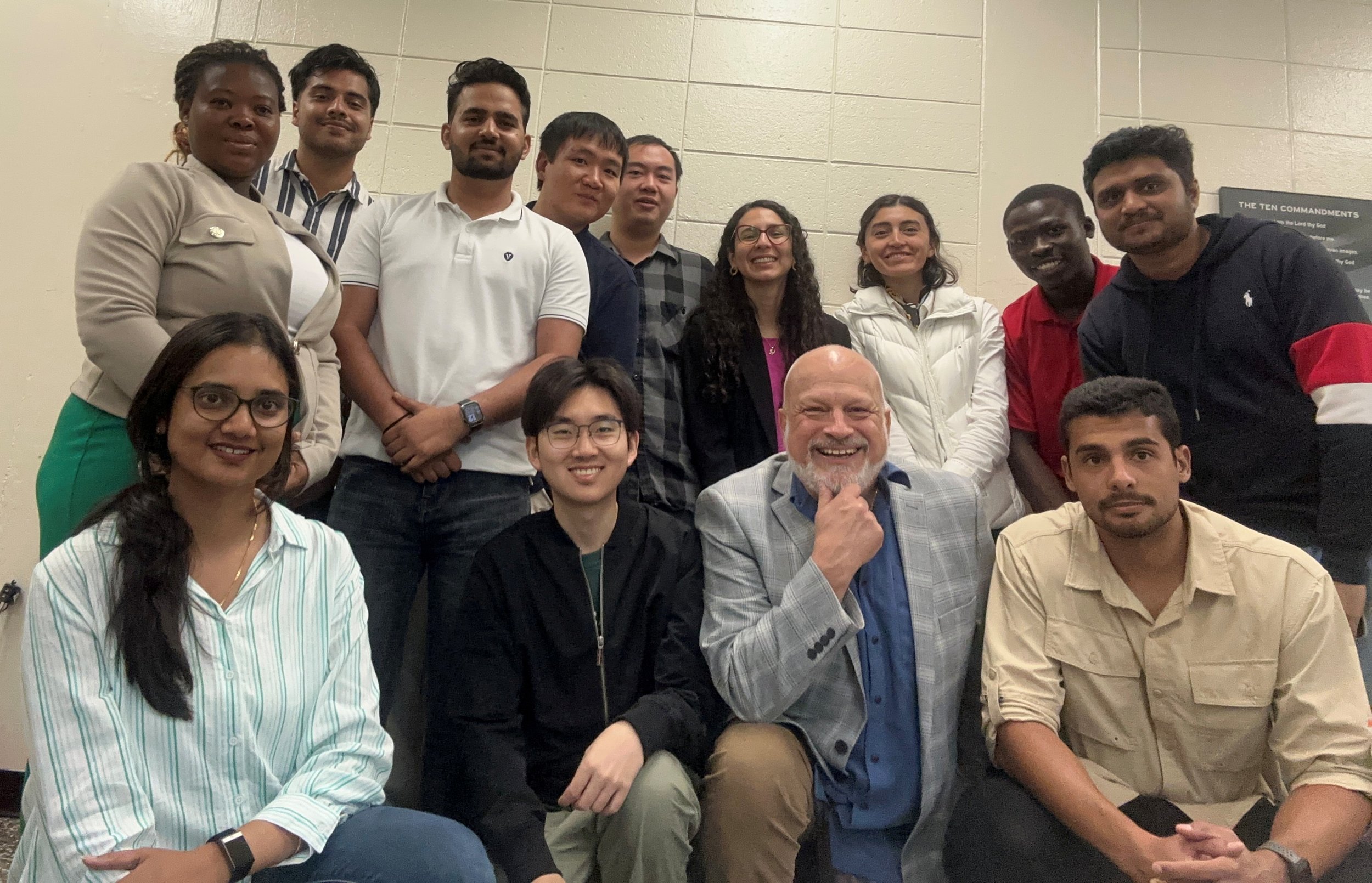 A group of diverse people (international students and Joseph Poniatowski) smiling and posing for a photo in a classroom at the University of Arkansas, with a beige wall in the background.