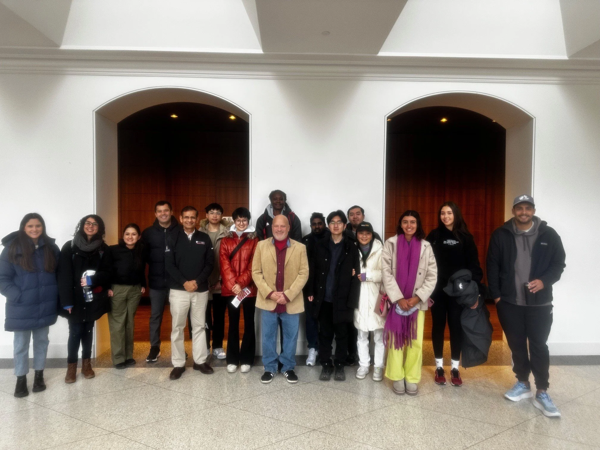 Group of international students and Joseph Poniatowski standing together indoors, smiling, in front of large wooden doors in a well-lit area, before the MLK day presentation by Little Rock Nine member, Minnijean Brown Trickey.