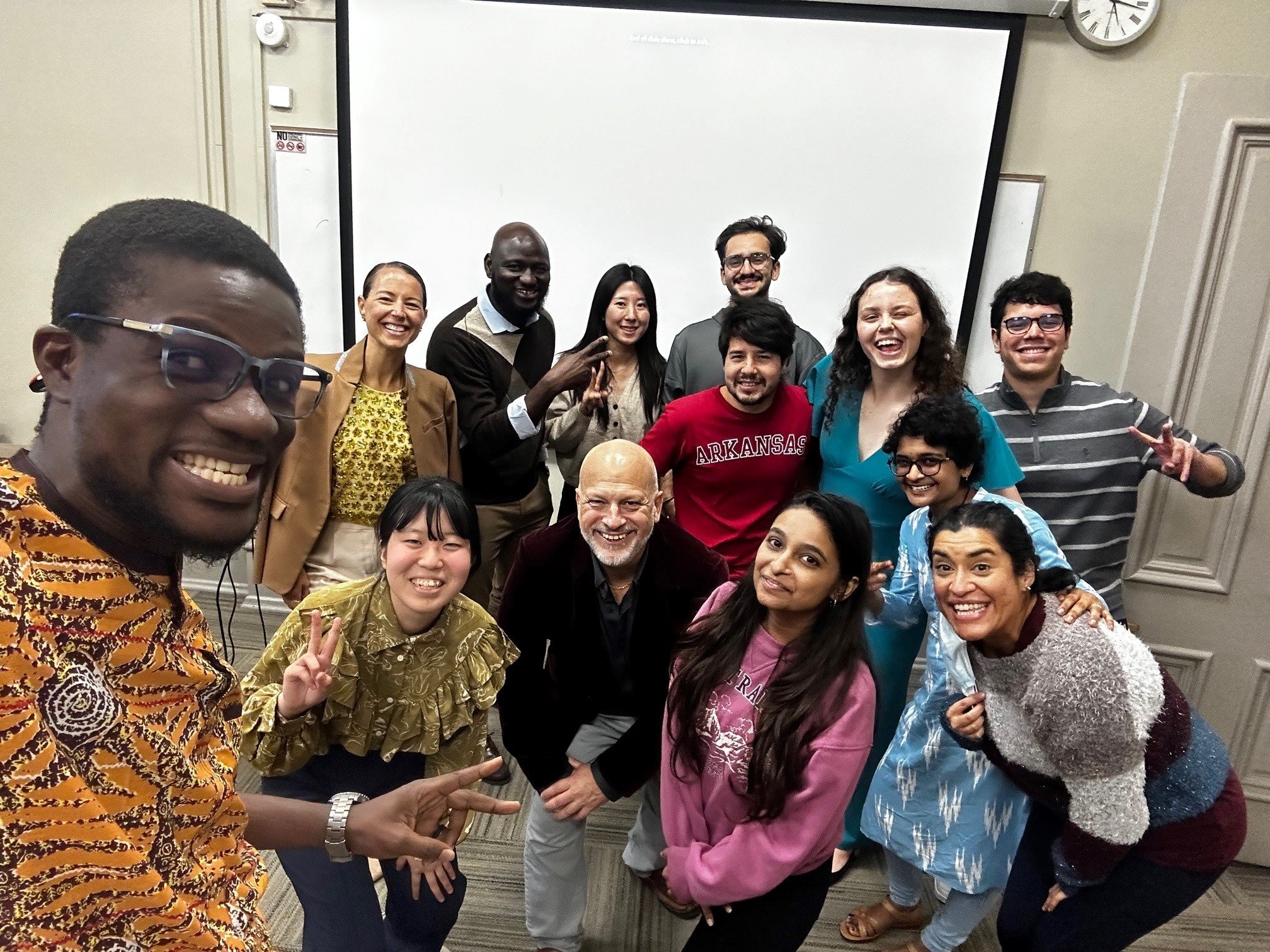 A group of diverse people (international students) and Joseph Poniatowski are smiling and posing for a photo in a room with a whiteboard and a clock on the wall in a classroom at the University of Arkansas.