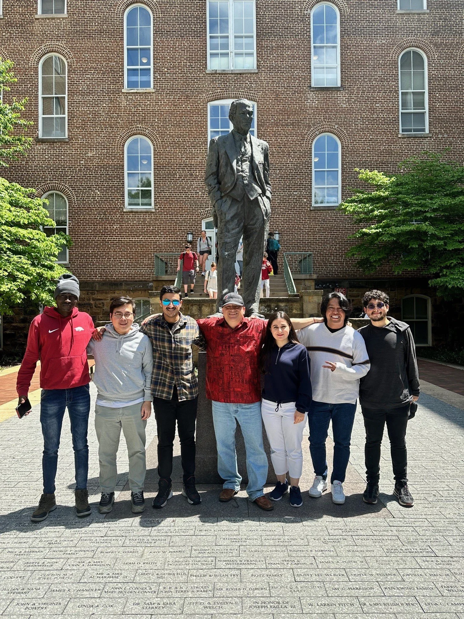 A group of eight international students along with Joseph Poniatowski standing in front of a memorial with a statue of William Fulbright on a brick building background. The people are smiling and posing for a photo, some have arms around each other
